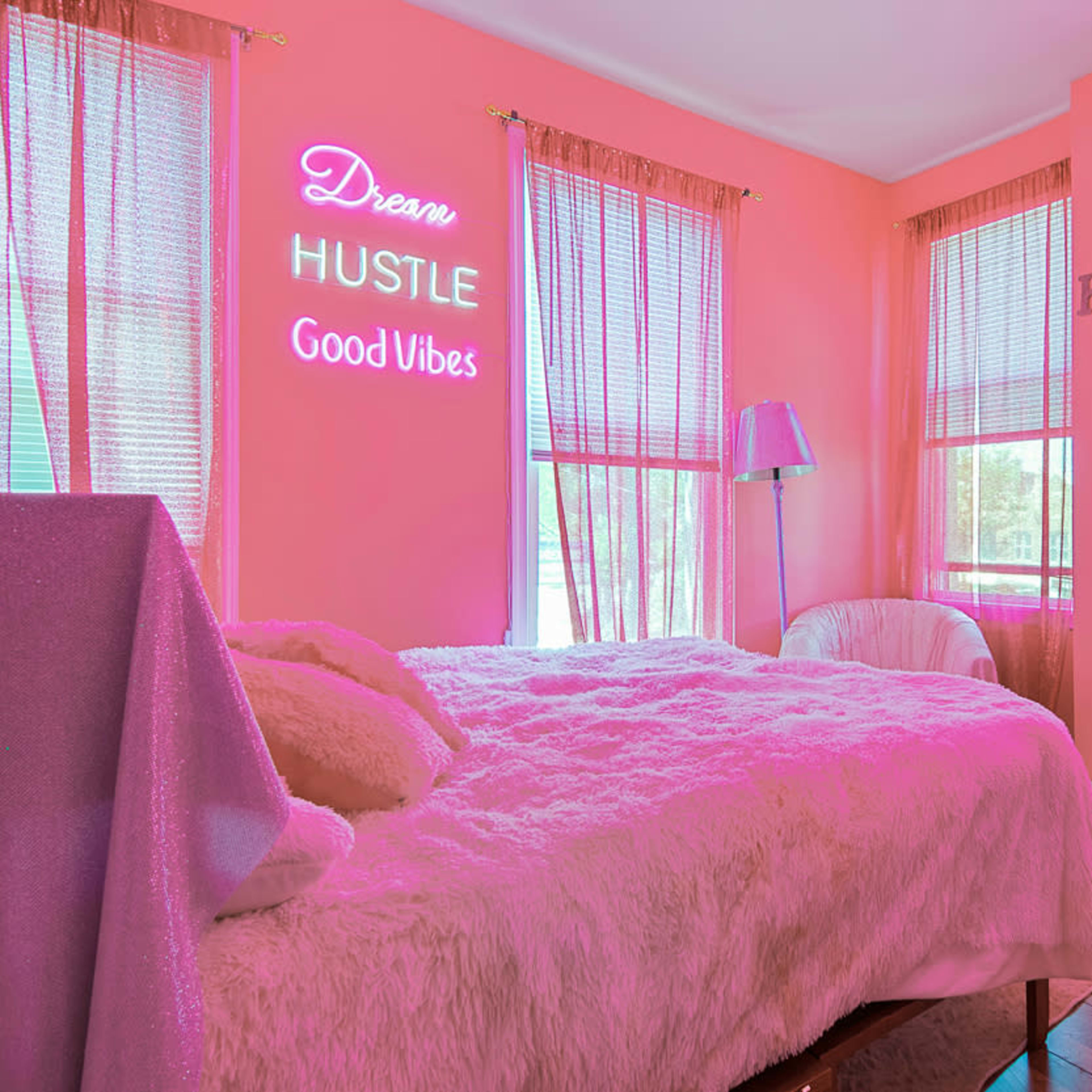 A brightly colored pink bedroom featuring a bed with a faux fur blanket, neon signs on the walls, and large windows letting in natural light.