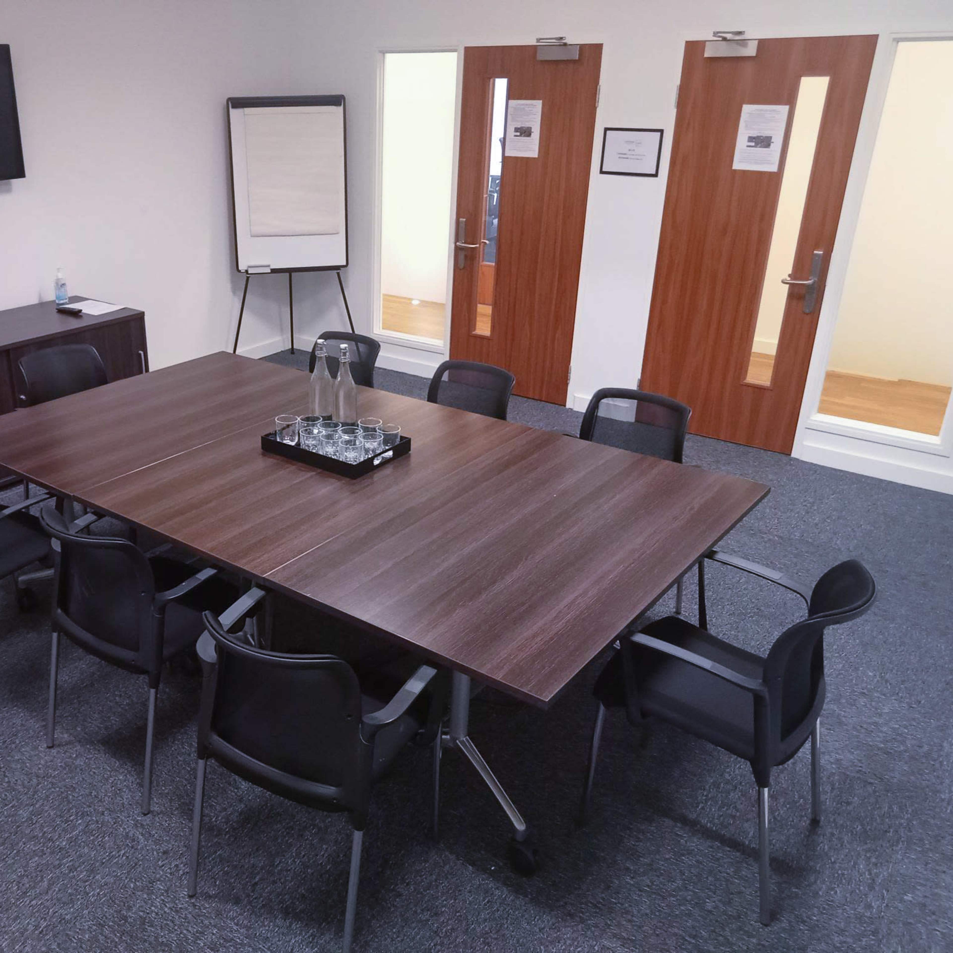 A meeting room features a large table surrounded by black chairs, a television mounted on the wall, and two doors in the background.