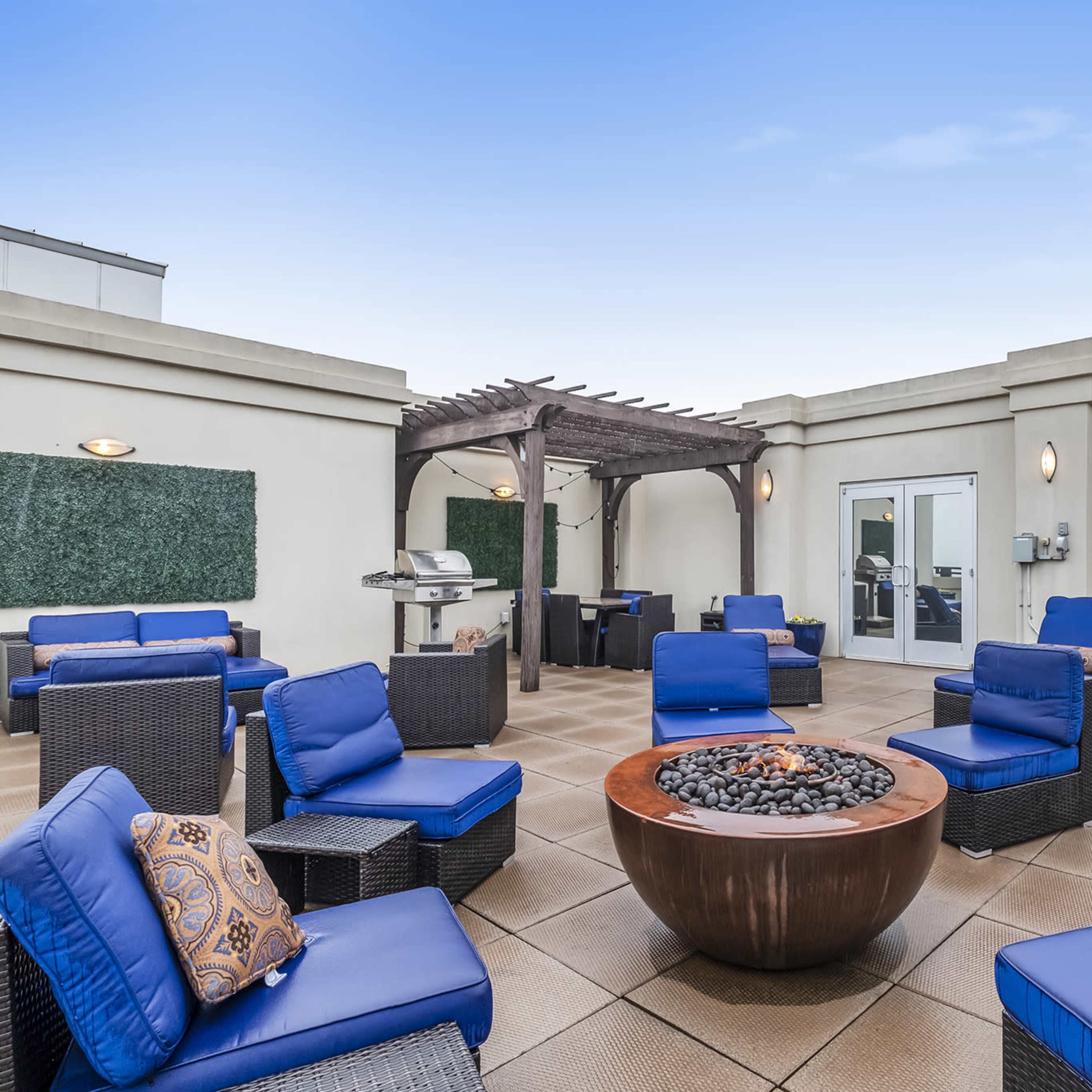 A rooftop lounge area with blue seating, a central fire pit, a grill station, and doors leading to an indoor space.