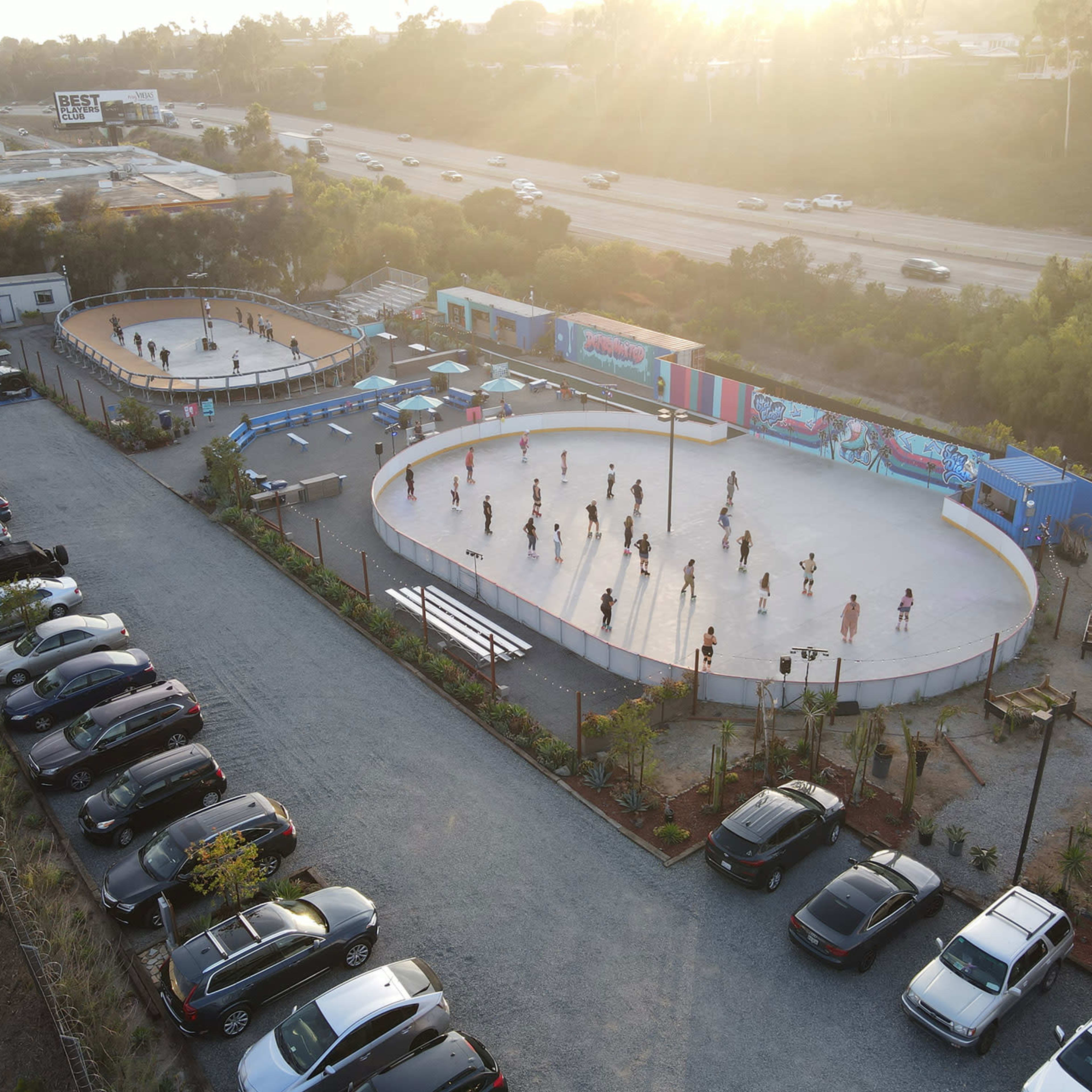 Two outdoor skating rinks with people skating, surrounded by cars parked in a gravel lot and bordered by greenery, under a sunset.