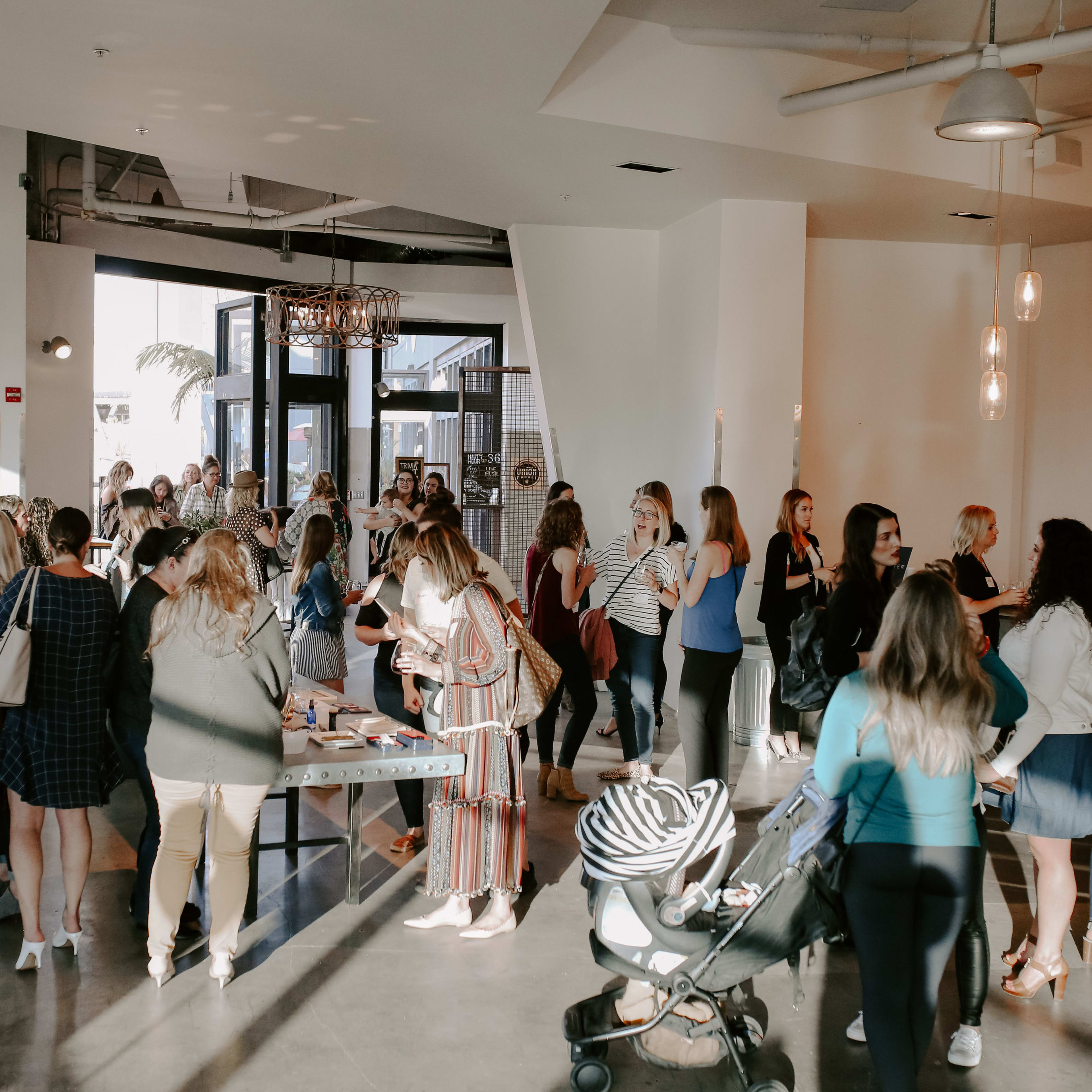A large group of people gathers in a modern indoor space, engaging in conversation and browsing various tables.