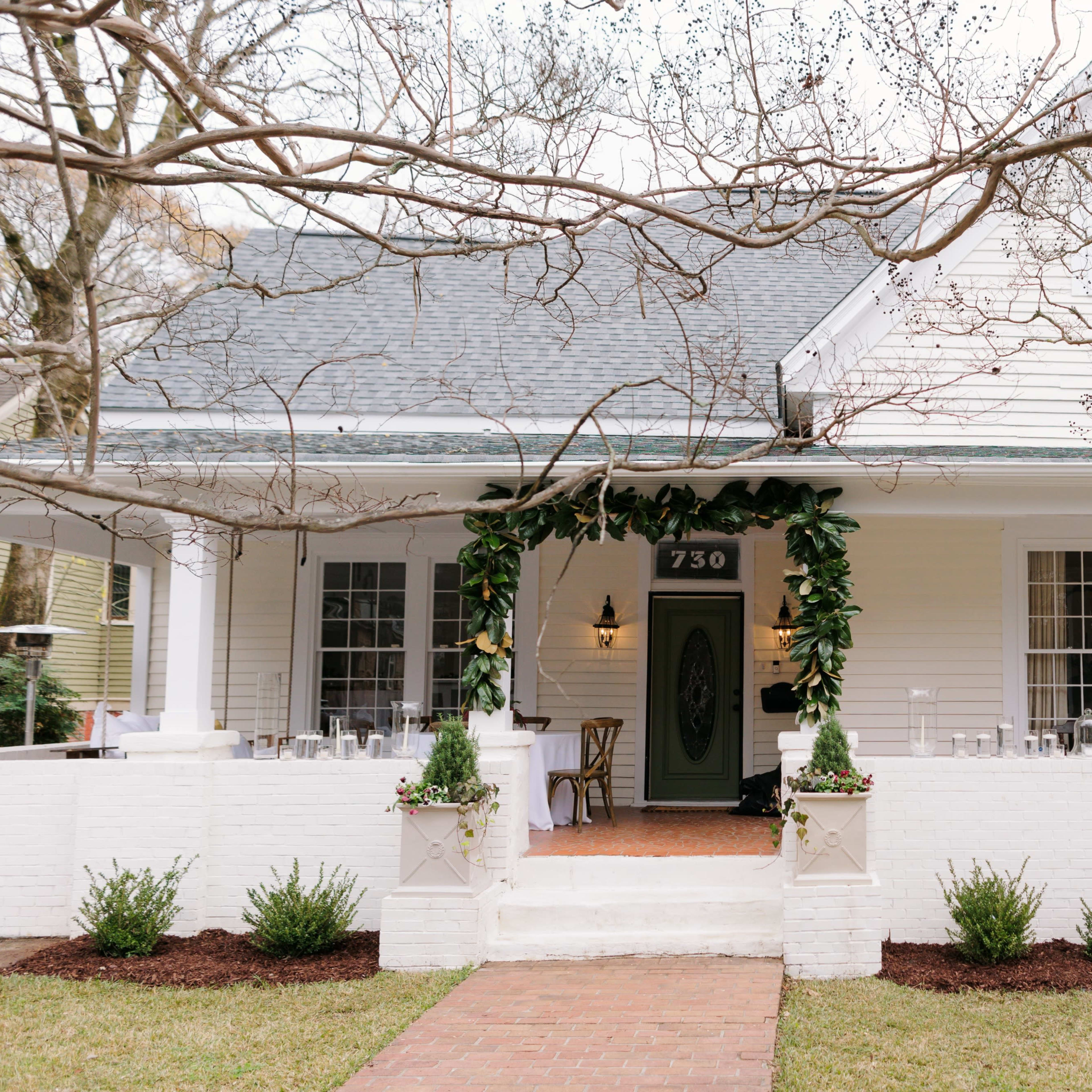 A white, single-story house with a covered porch is adorned with greenery around the entrance, featuring a brick pathway and neatly landscaped shrubs.