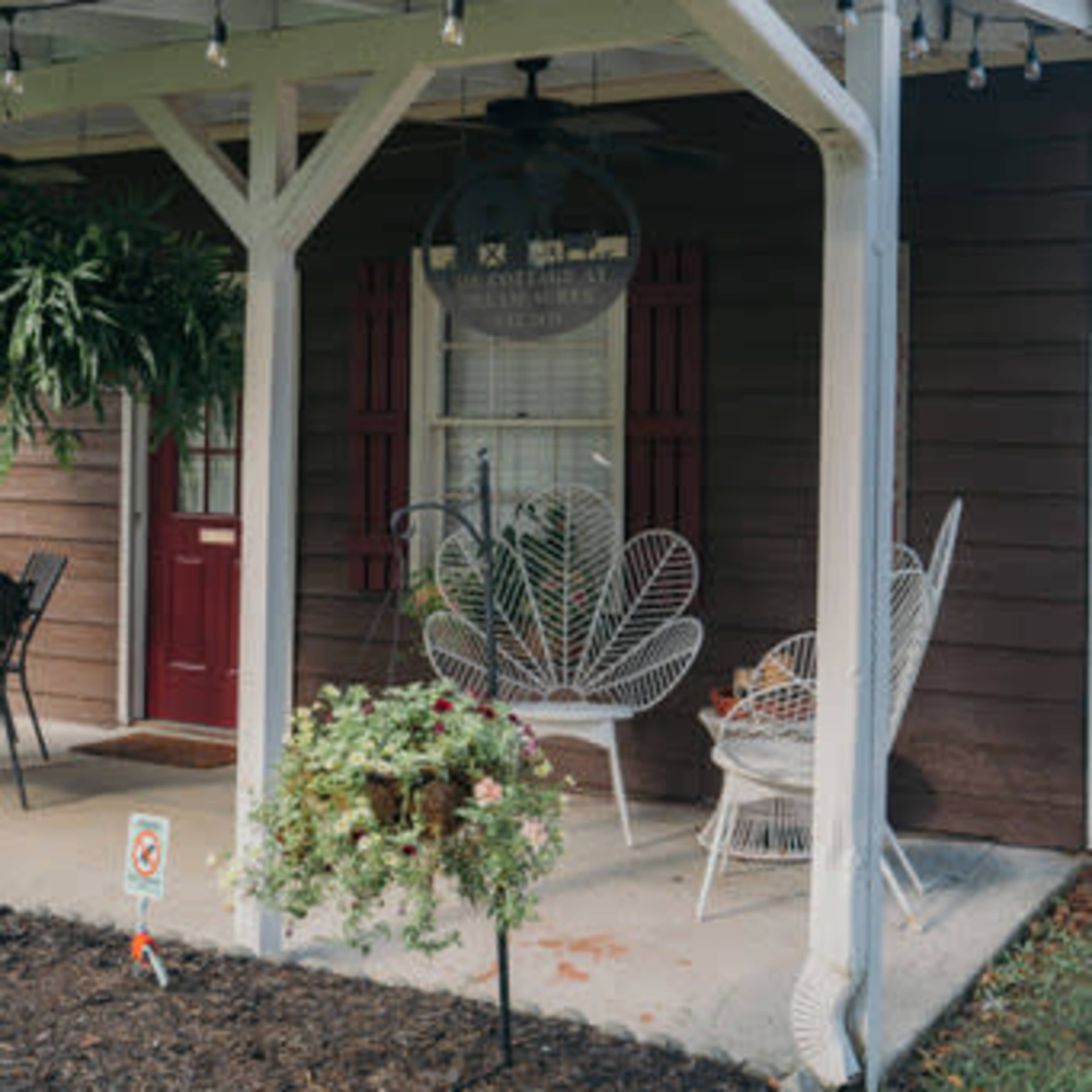 A porch with a ceiling fan features two white wicker chairs and hanging plants, set against a brown wooden house with red shutters.