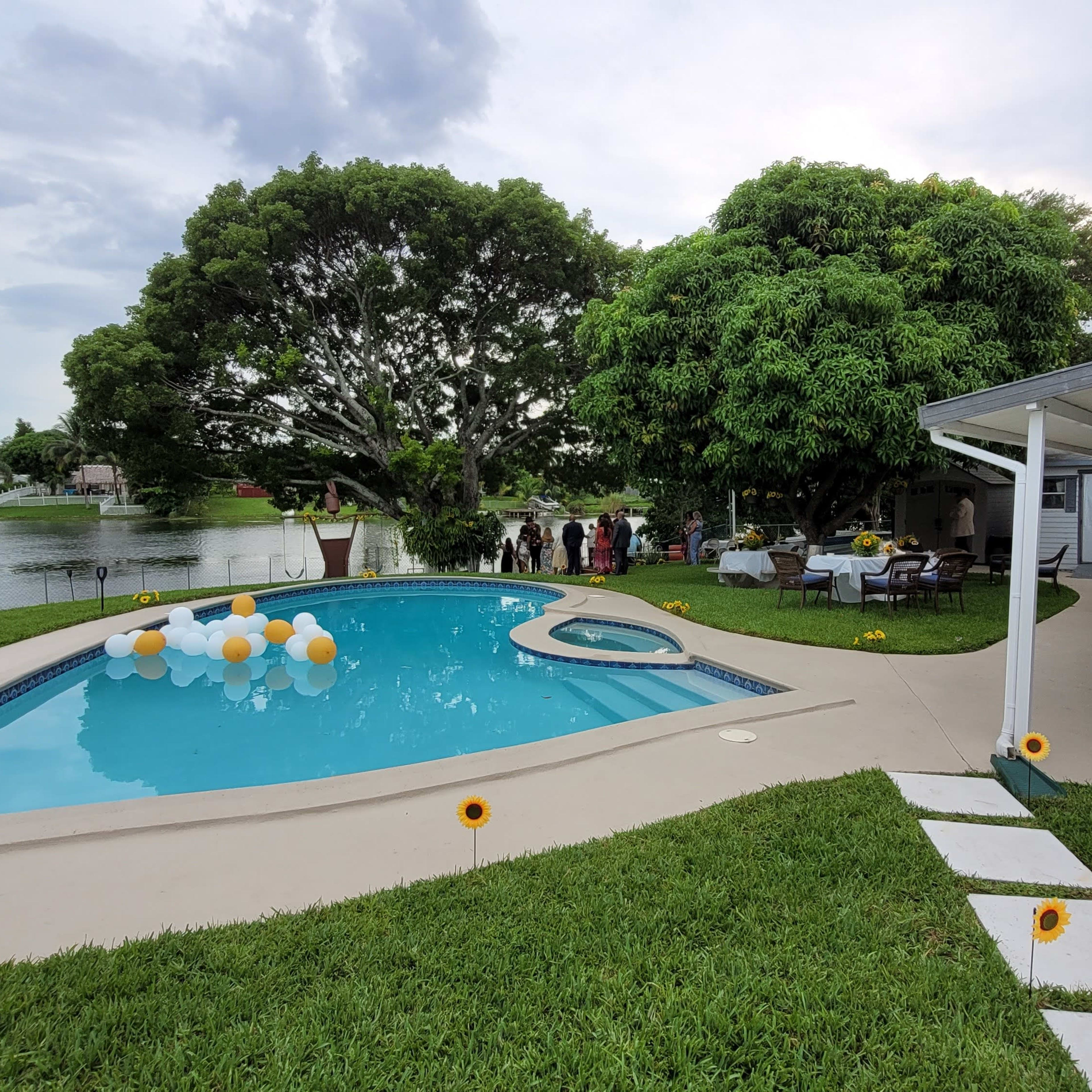 The scene features a backyard pool area adorned with balloons and seating arrangements, alongside a scenic lake and lush greenery.