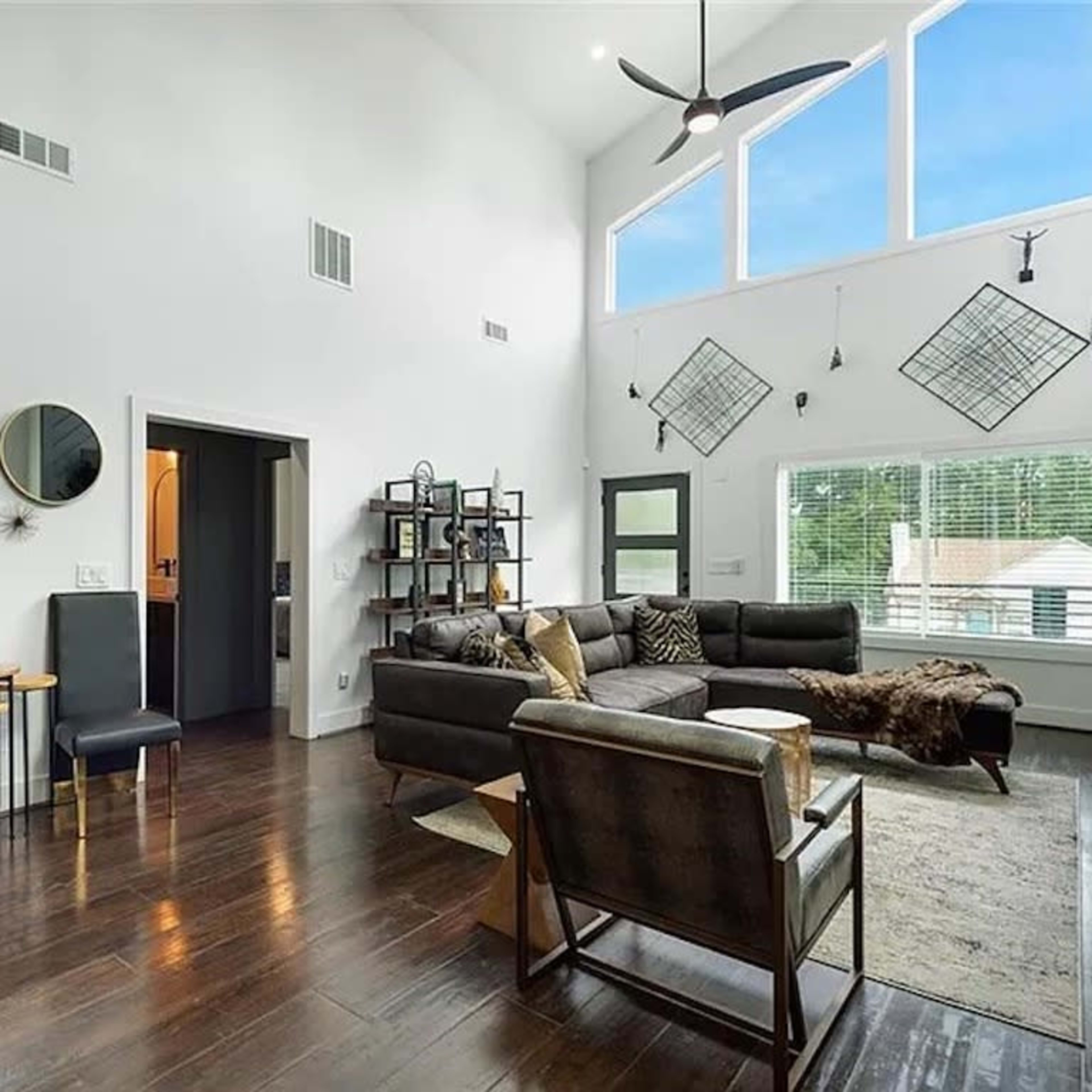 A modern living room features large windows, a sectional sofa, a shelving unit, and decorative mirrors on the walls.