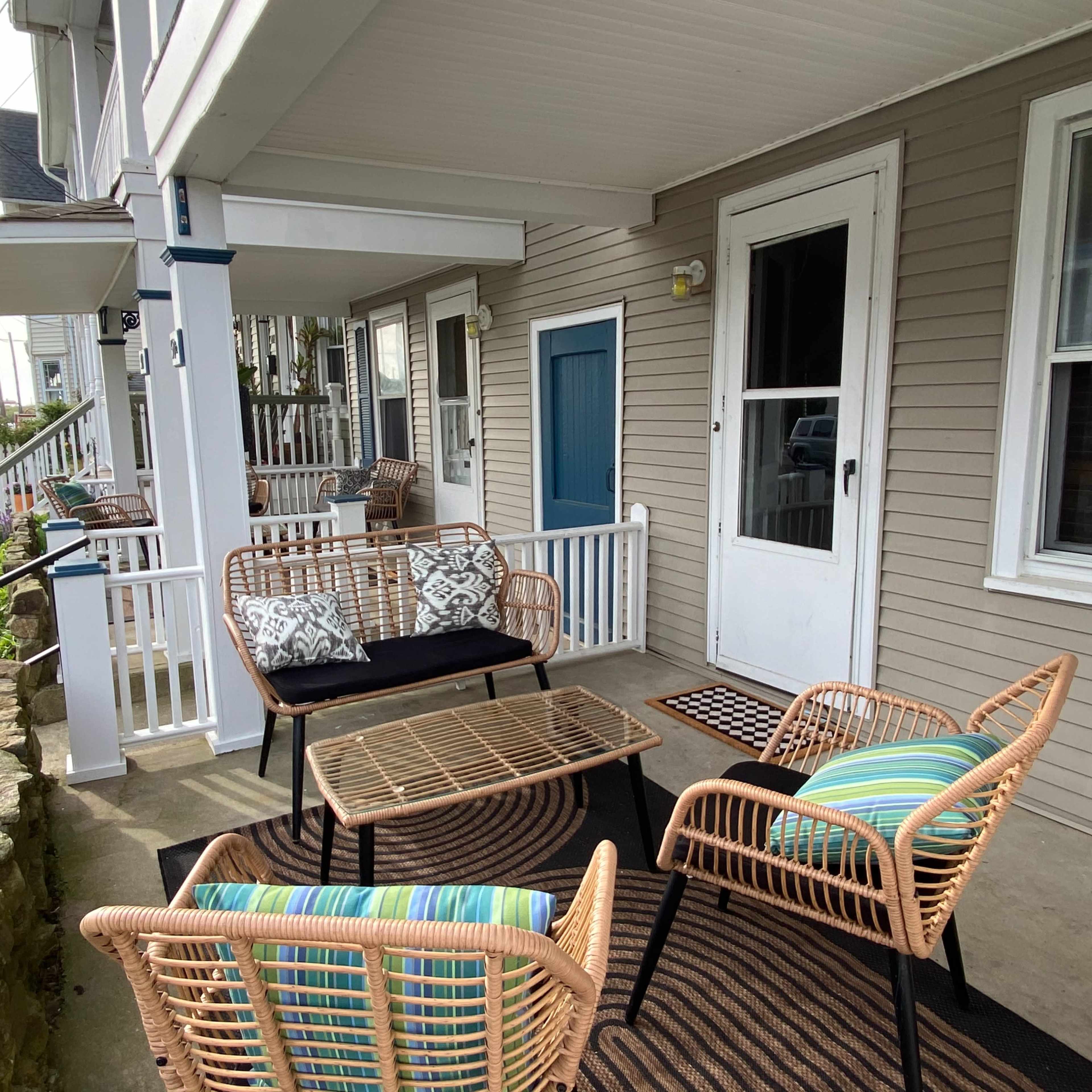A covered porch with a seating area featuring wicker chairs and a table, alongside a front door and a pathway lined with greenery.