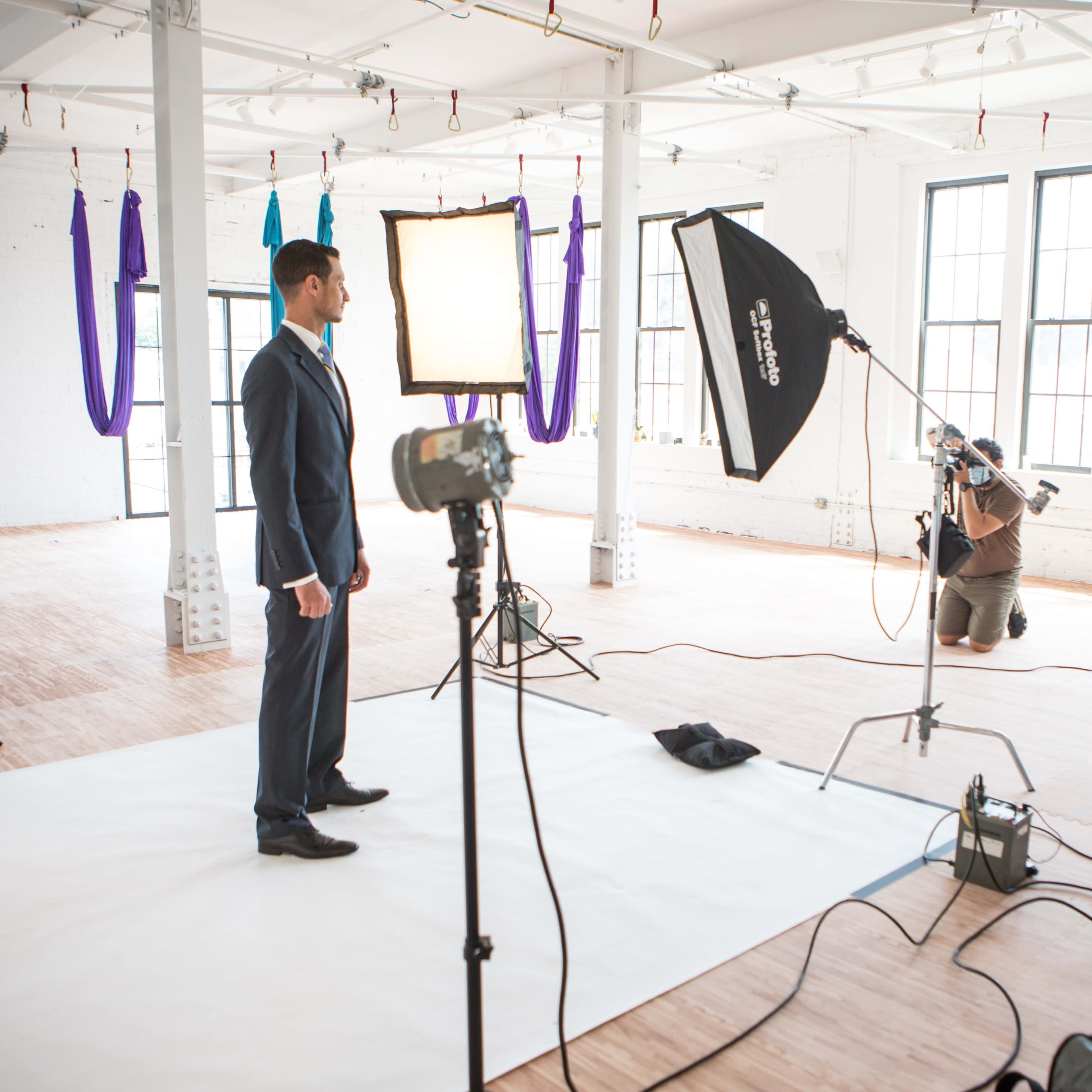 A man in a suit stands on a white backdrop, while a photographer kneels behind a camera and softbox in a well-lit studio.