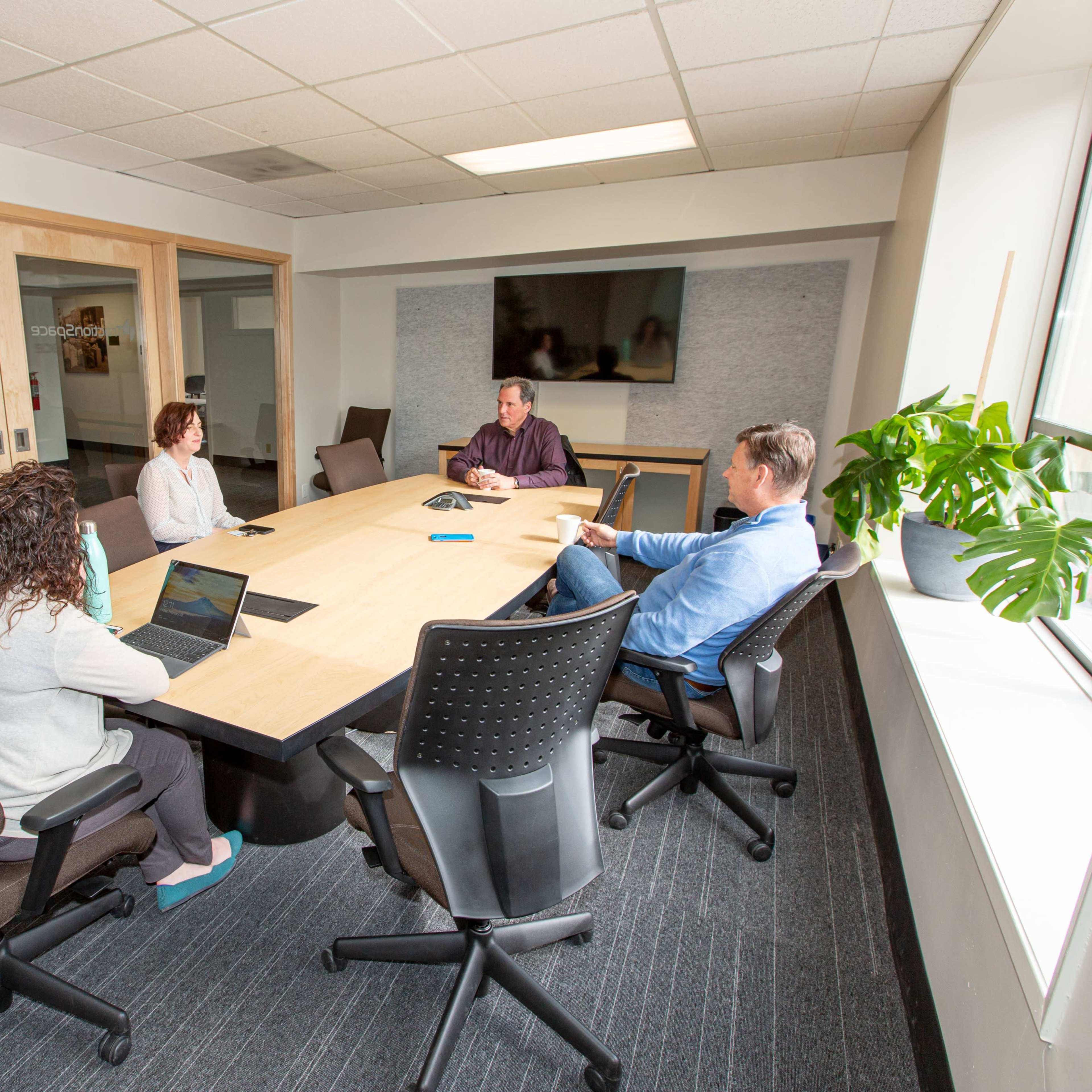A group of four people are seated around a rectangular conference table in a modern meeting room, with laptops and documents in front of them.
