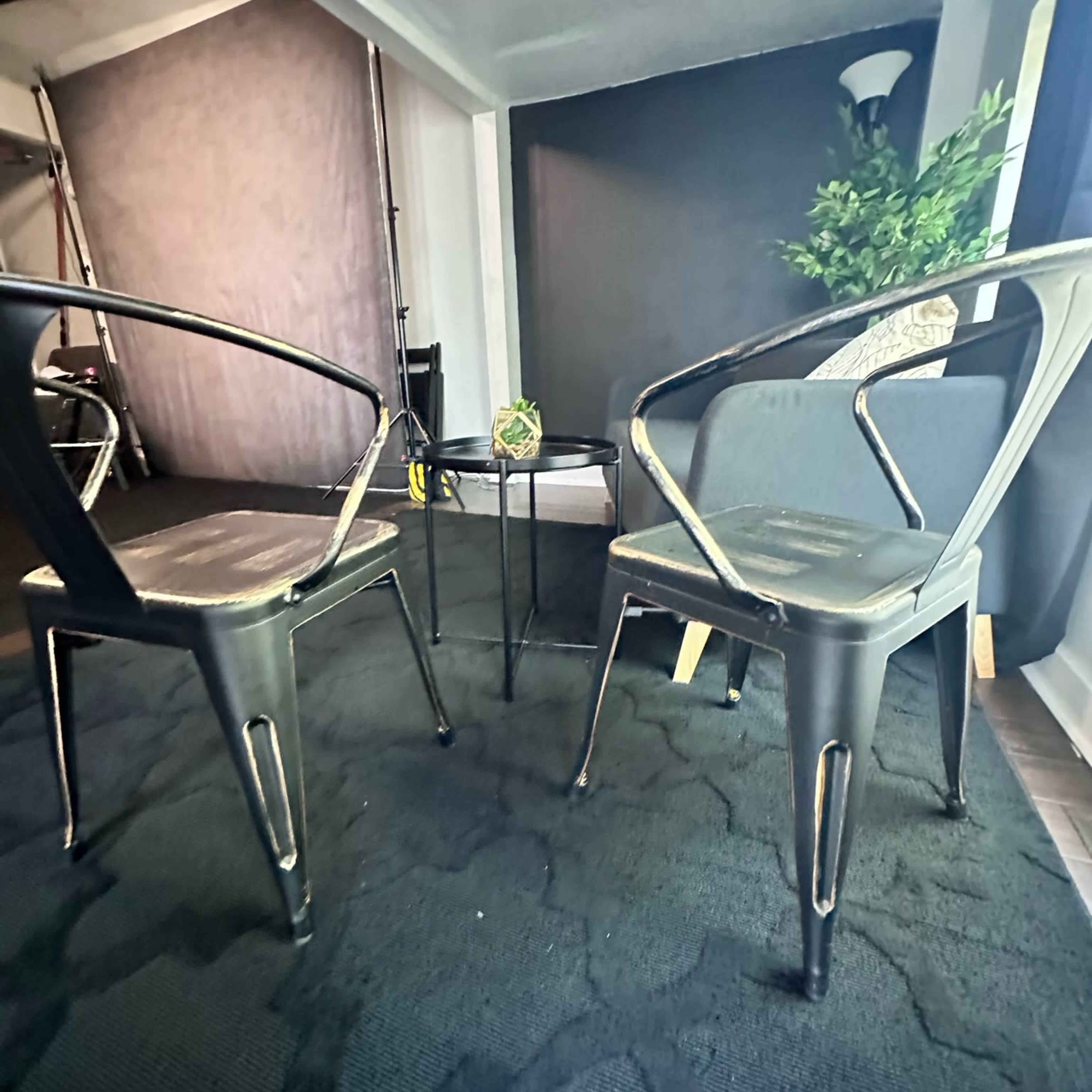 Two metal chairs face each other across a small round table in a room with dark walls and a plant in the background.
