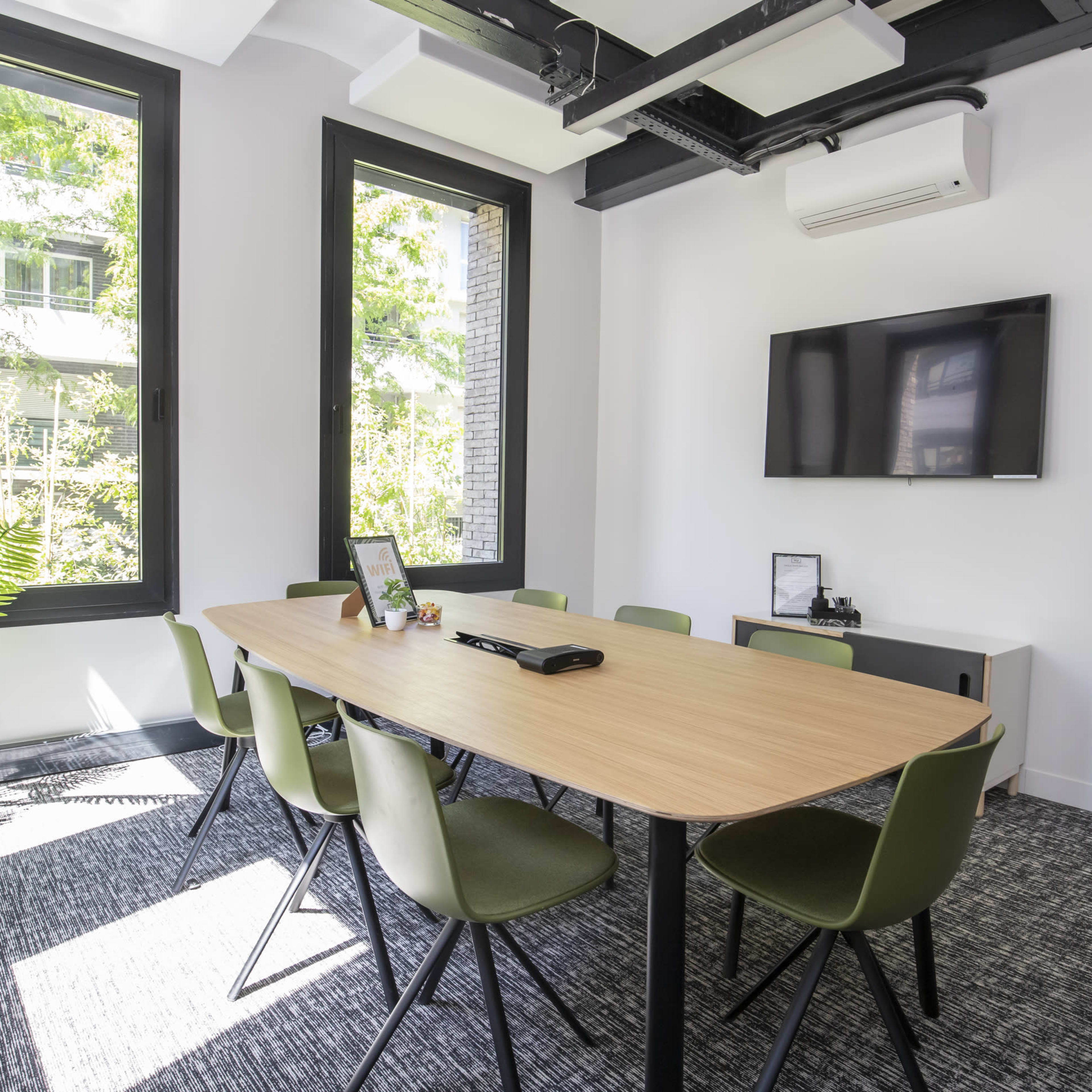 A meeting room features a large wooden table surrounded by green chairs, with a television mounted on the wall and large windows allowing natural light to enter.