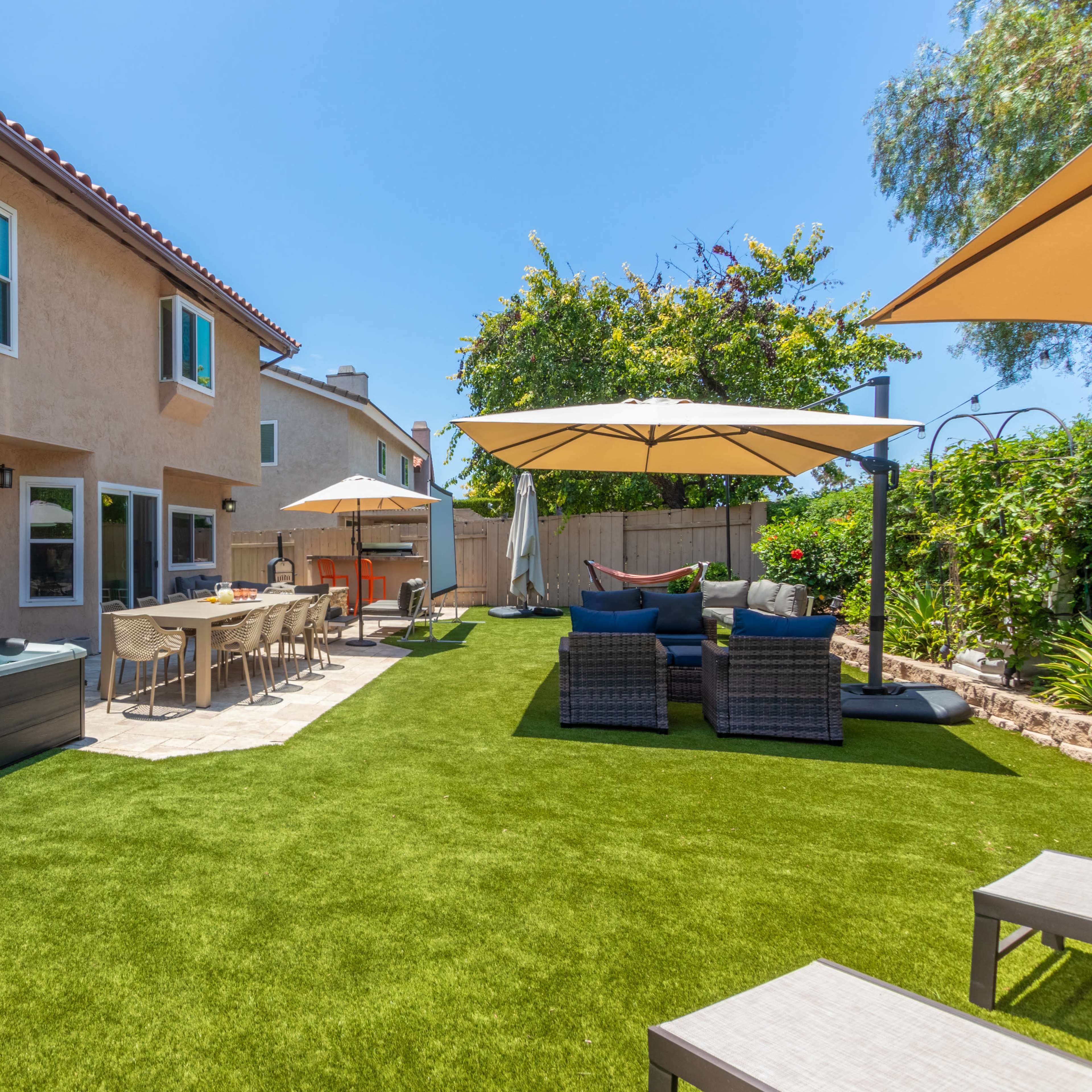A backyard with a hot tub, outdoor seating furniture, dining table, and umbrellas set on green artificial grass.