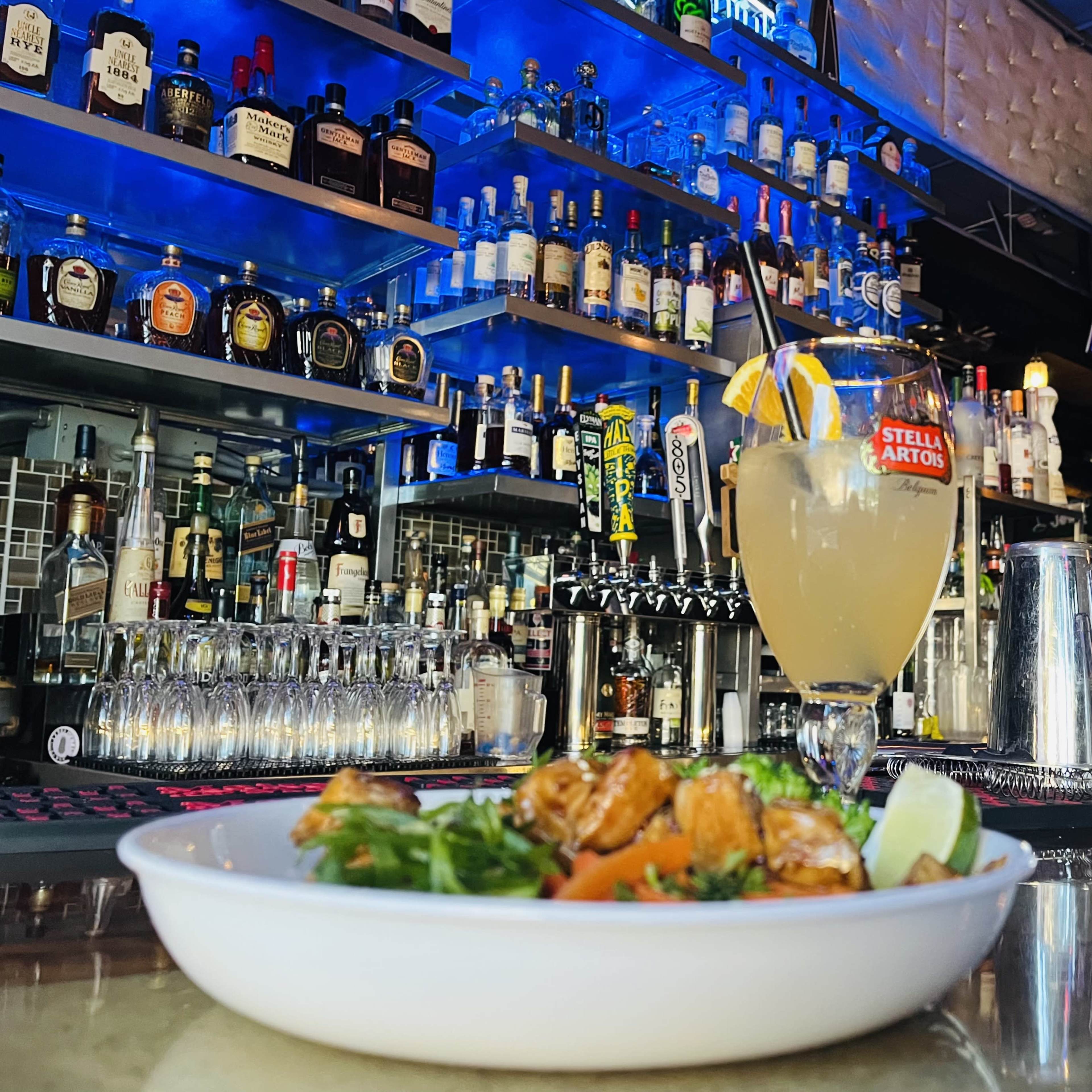 A plate of food sits on the bar in front of a colorful cocktail, with a well-stocked shelf of liquor bottles and a television displaying a show in the background.