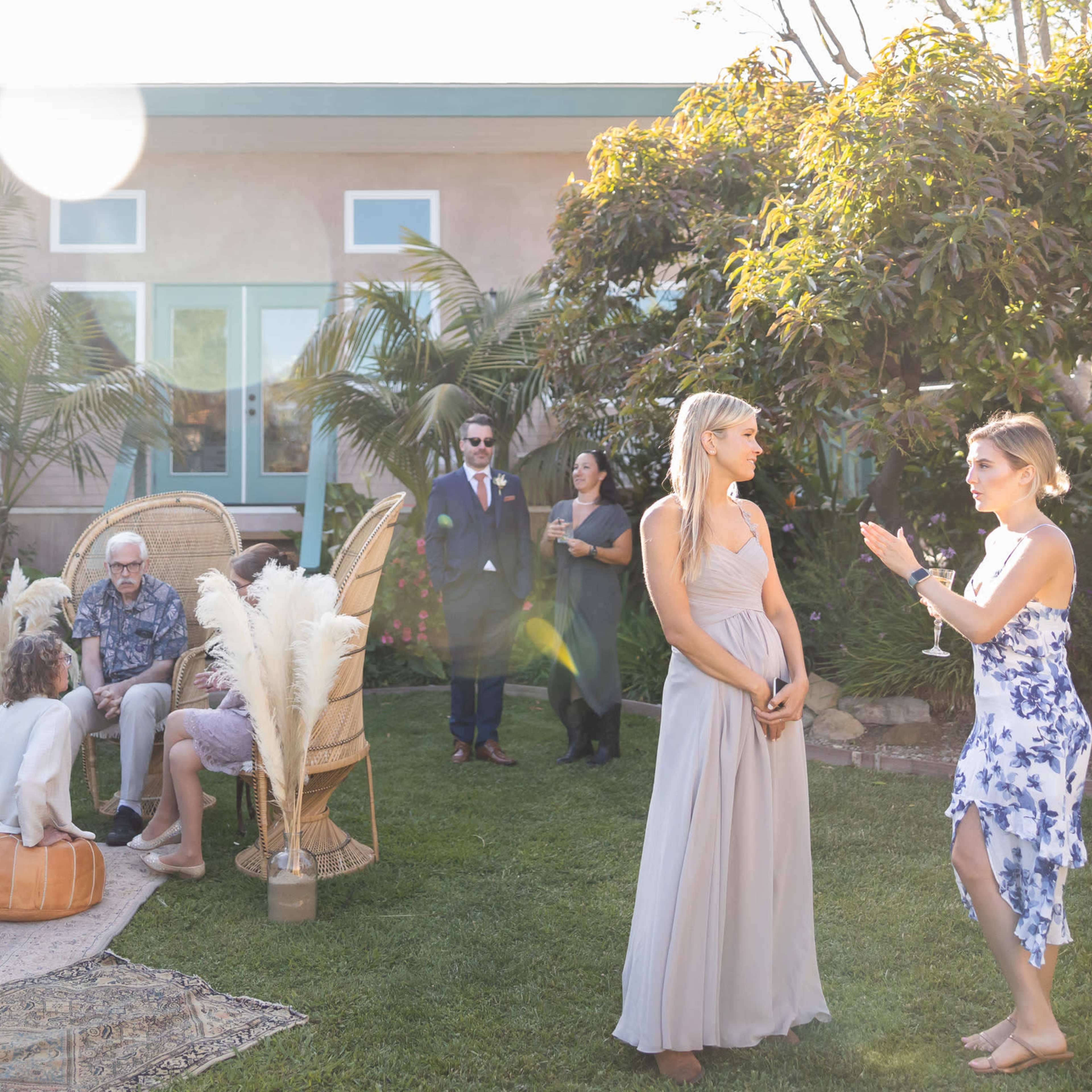 A group of people mingles outdoors in a garden setting, with decorative furniture and greenery surrounding them.