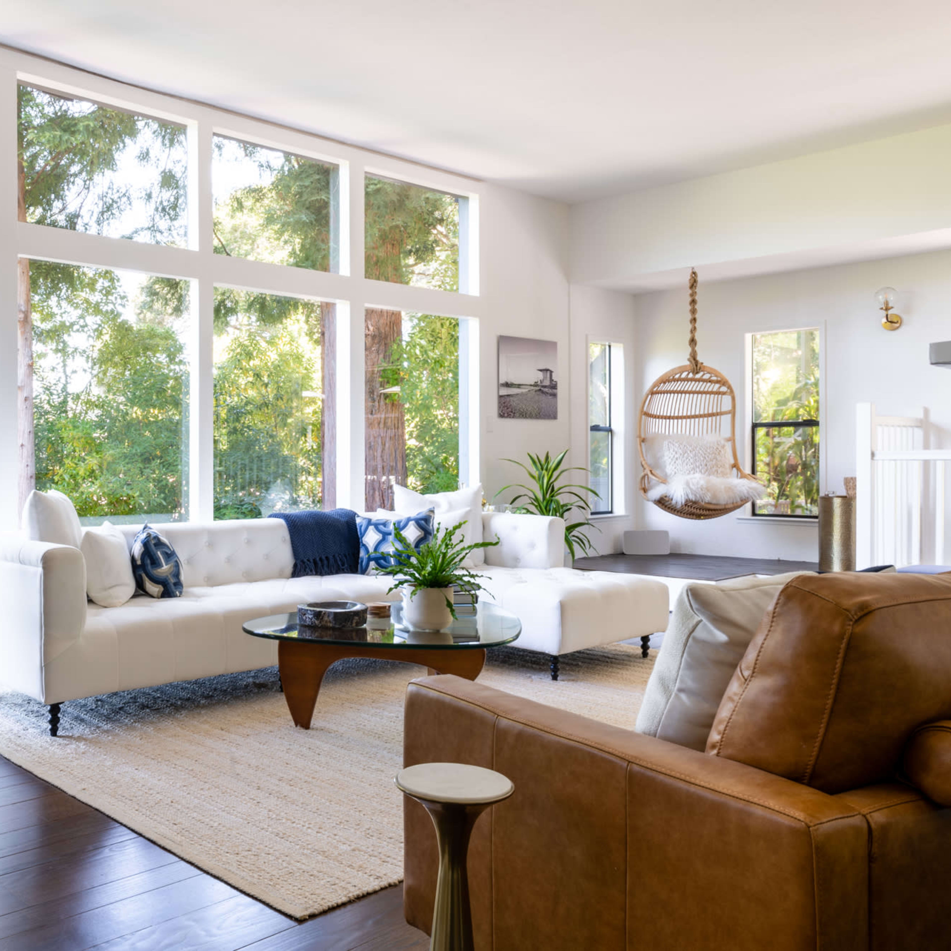 A bright living room with large windows showcasing greenery outside, featuring a white sofa, a glass coffee table, and a hanging chair.
