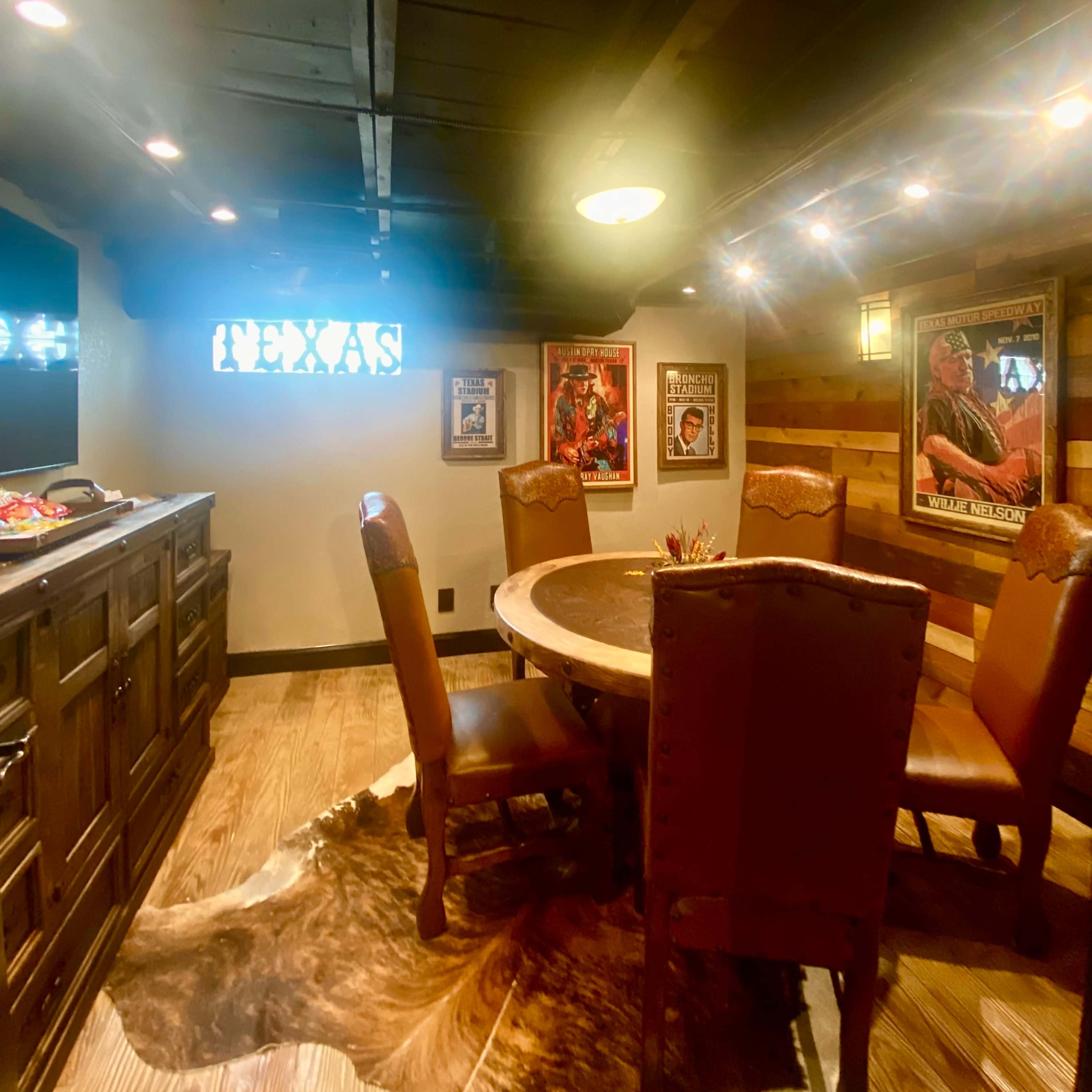 A rustic dining area features a round wooden table surrounded by upholstered chairs, with framed posters on the walls and wooden accents throughout.