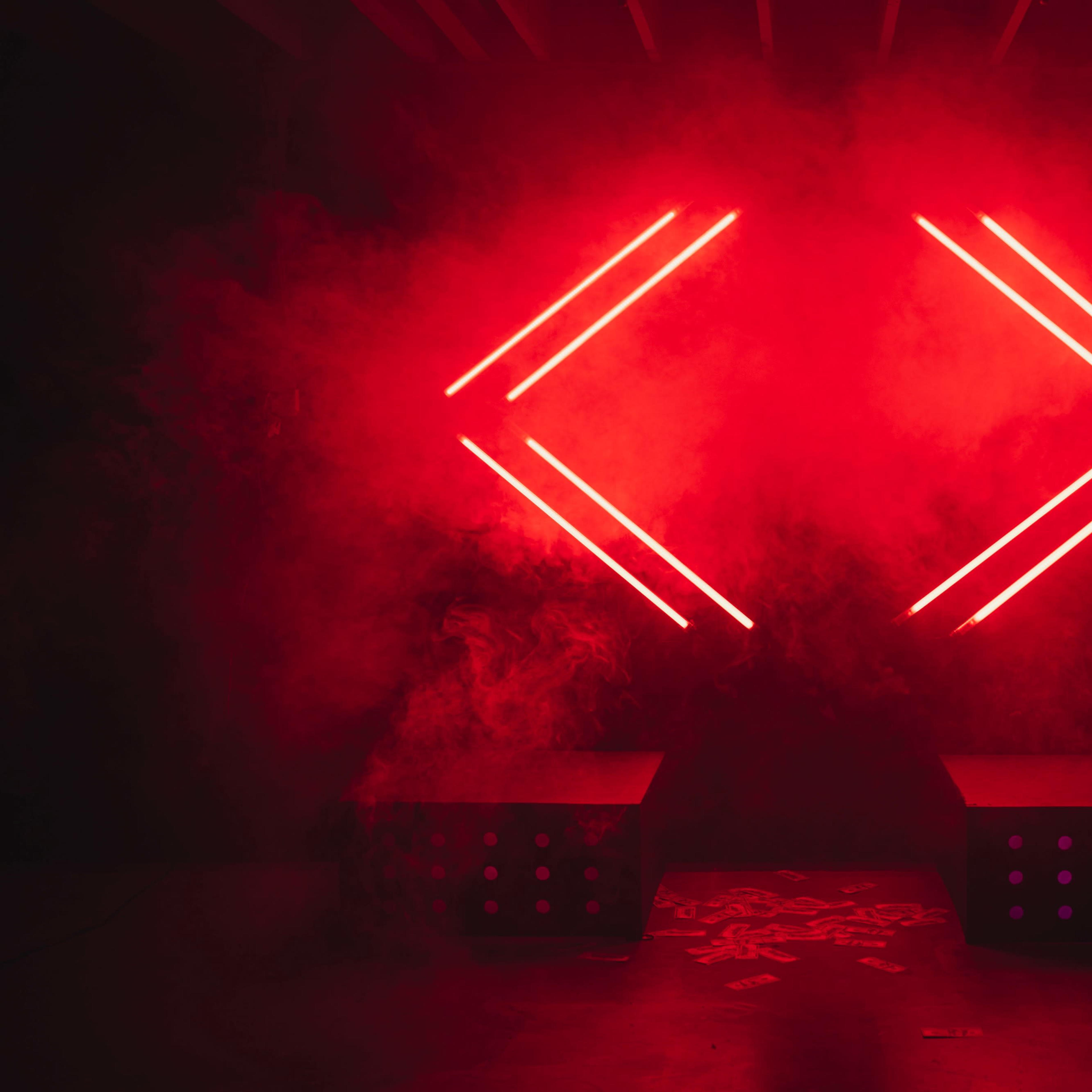Two black boxes with dotted patterns in front of a backdrop illuminated by two red neon arrows amidst a smoky atmosphere.