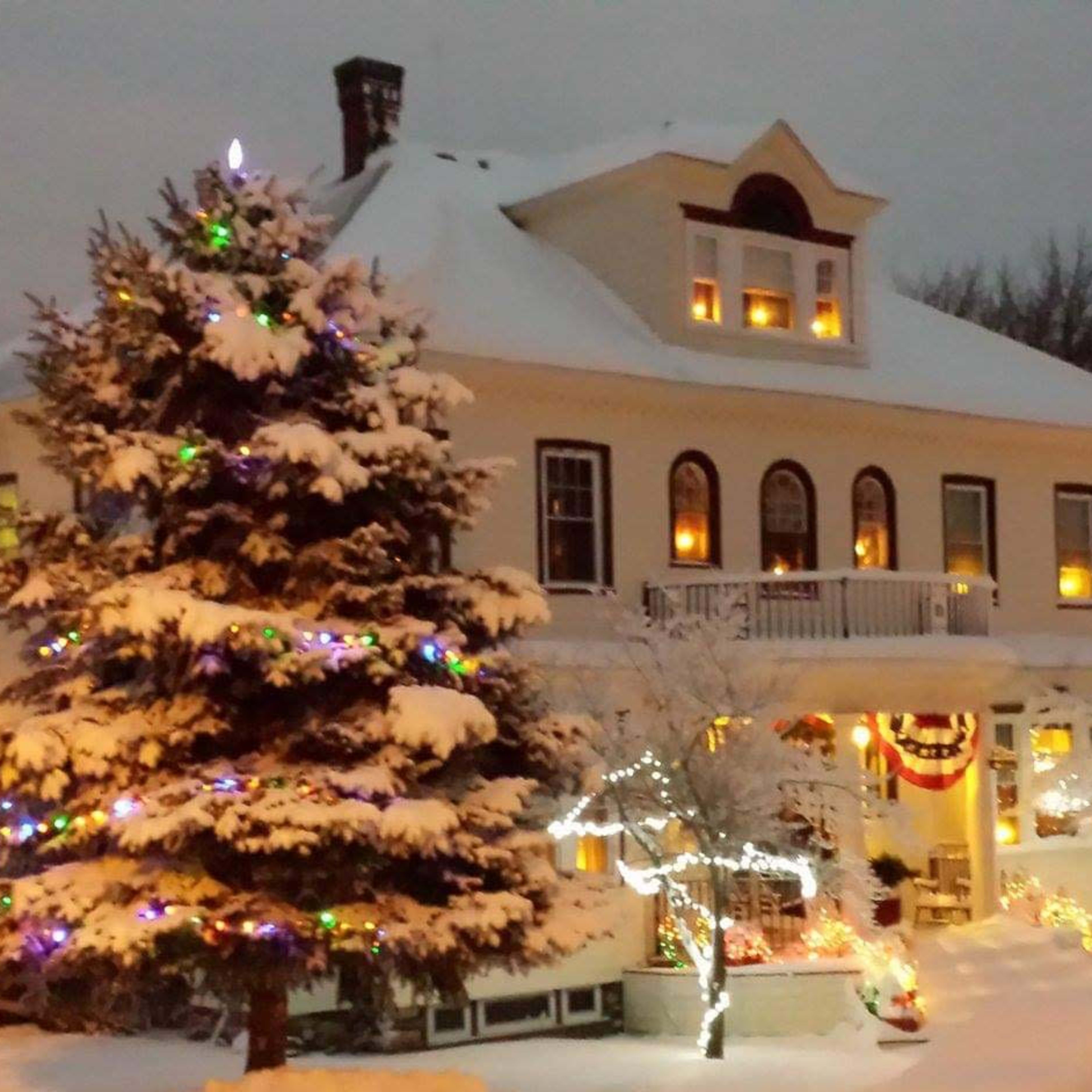 A two-story house with numerous windows illuminated, surrounded by snow and decorated with colorful holiday lights and a snow-covered tree in the foreground.
