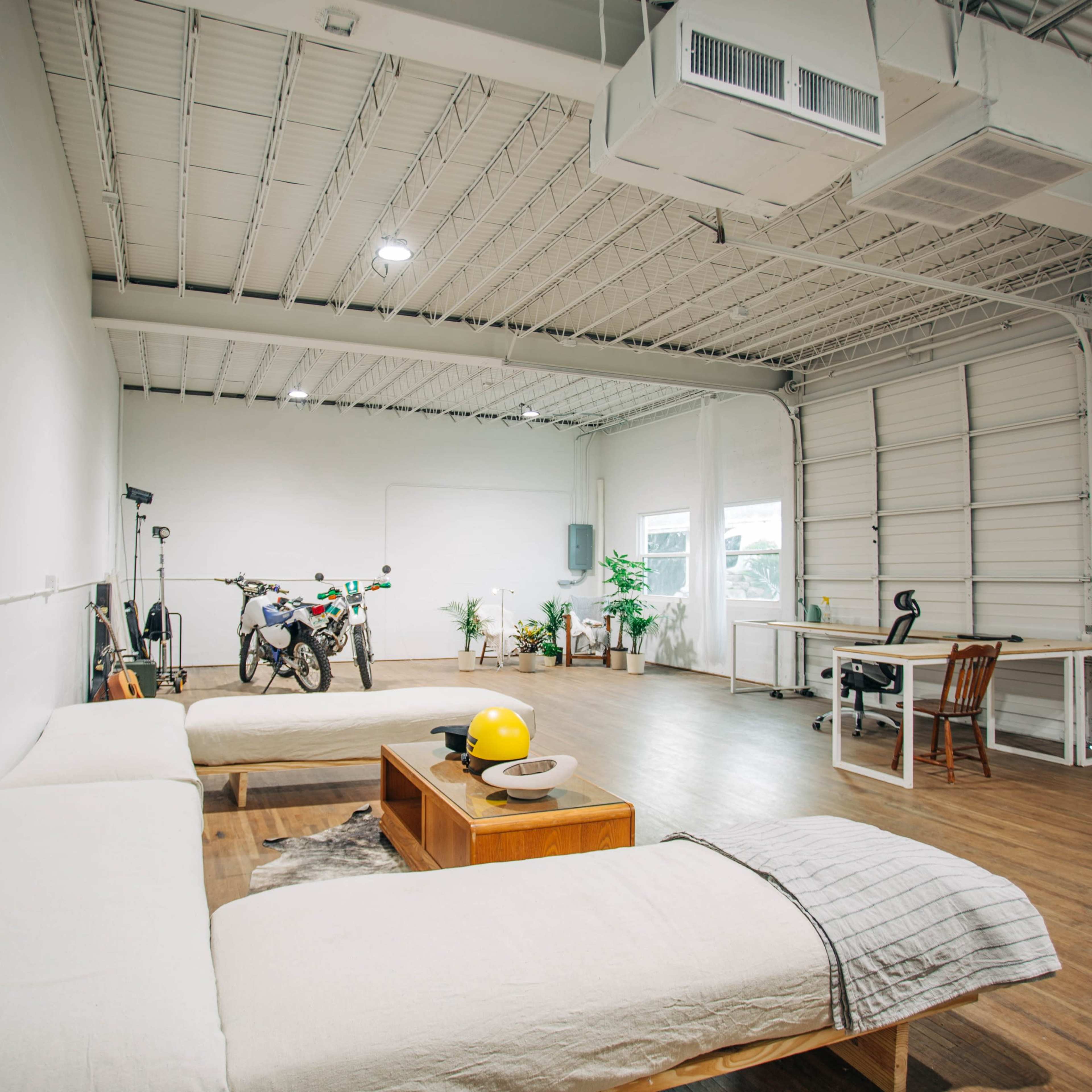 A spacious, minimalist room with a large couch, a wooden coffee table, a motorcycle, plants, and a work desk.
