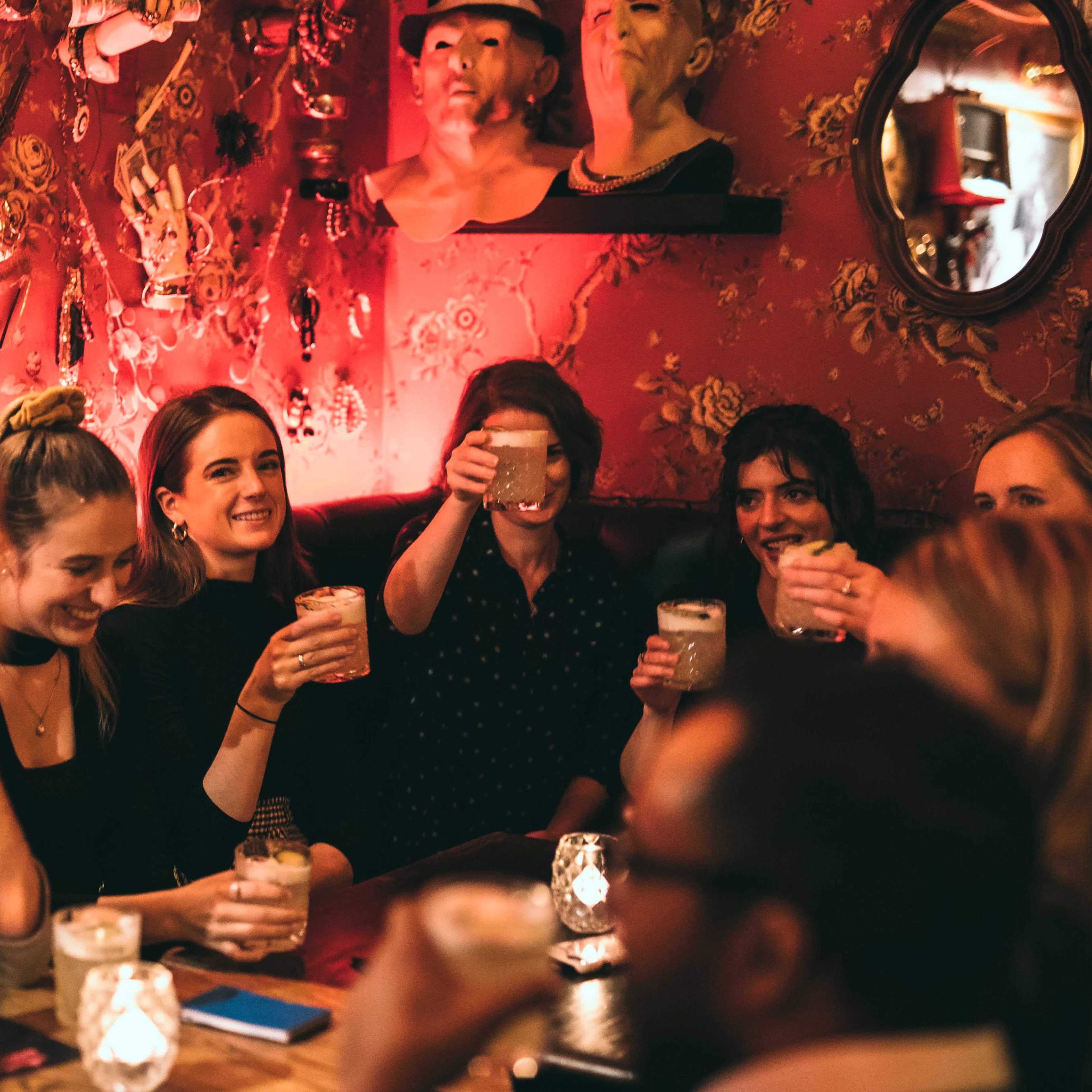 A group of people is raising their drinks in a dimly lit bar decorated with vintage clocks and masks.