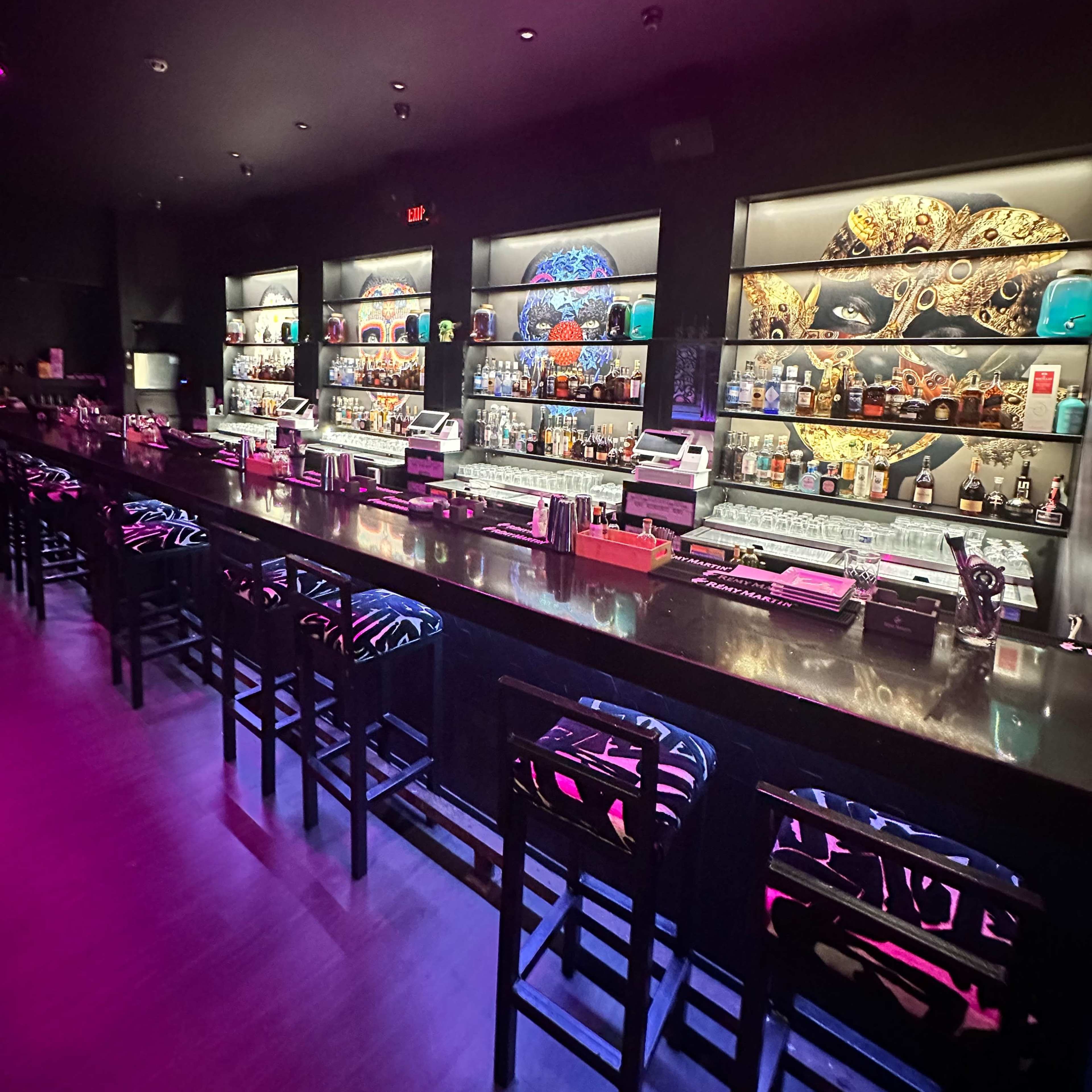 A dimly lit bar with a polished countertop and shelves displaying various bottles of alcohol against a colorful backdrop.