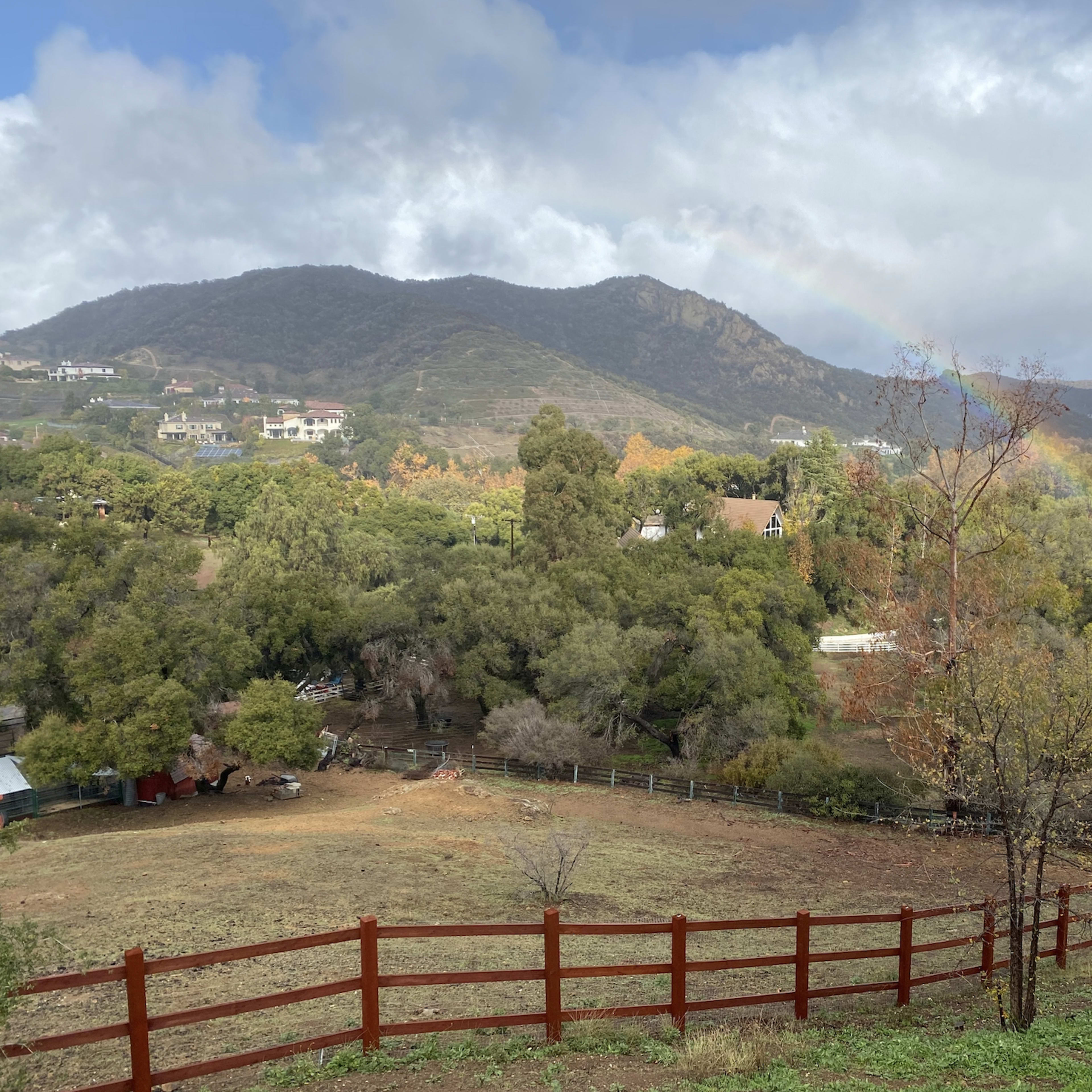 A wooden fence borders a grassy field with scattered trees and a rainbow arching over the distant mountains.