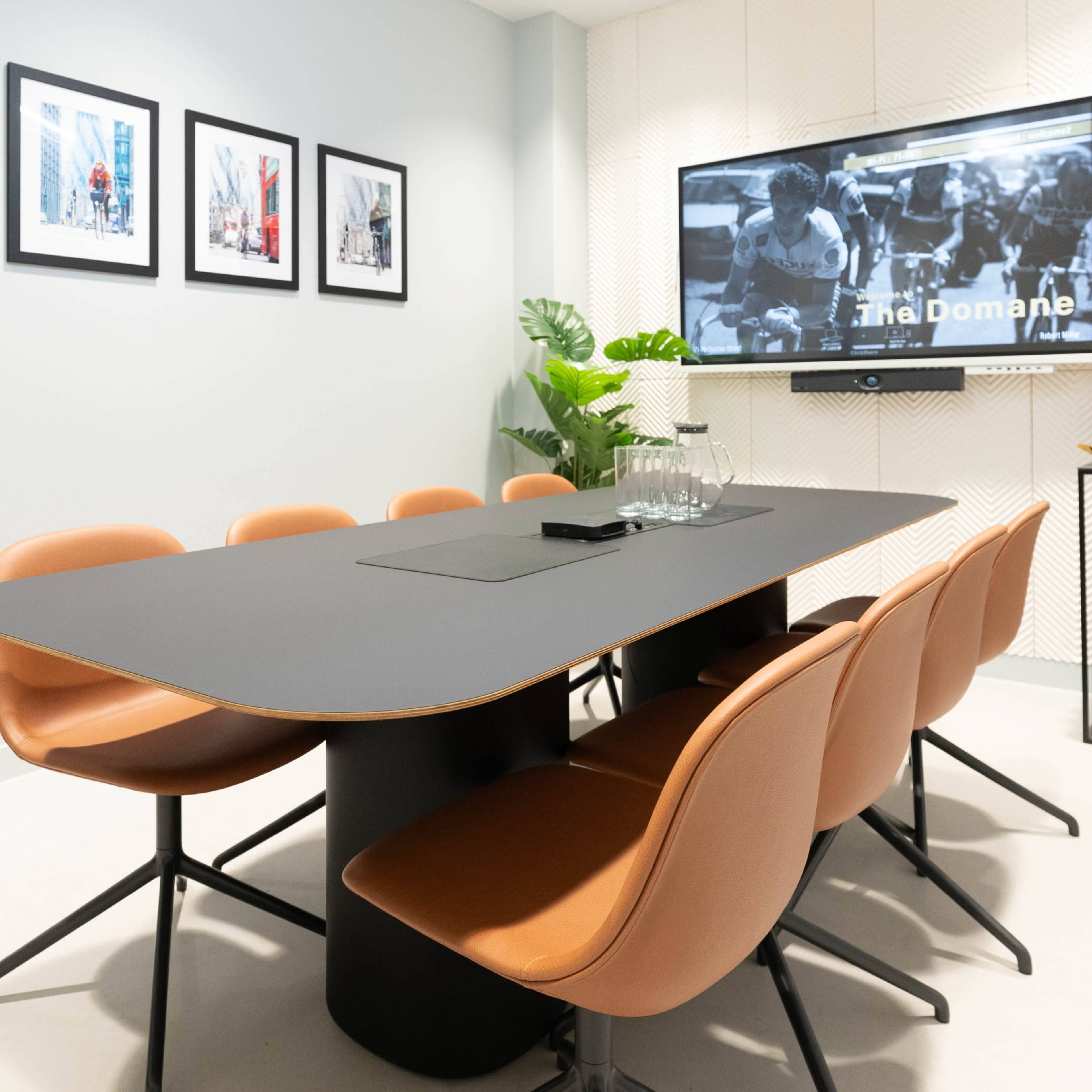 A modern conference room features a sleek black table surrounded by brown chairs, with framed artwork on the wall and a TV displaying a presentation.