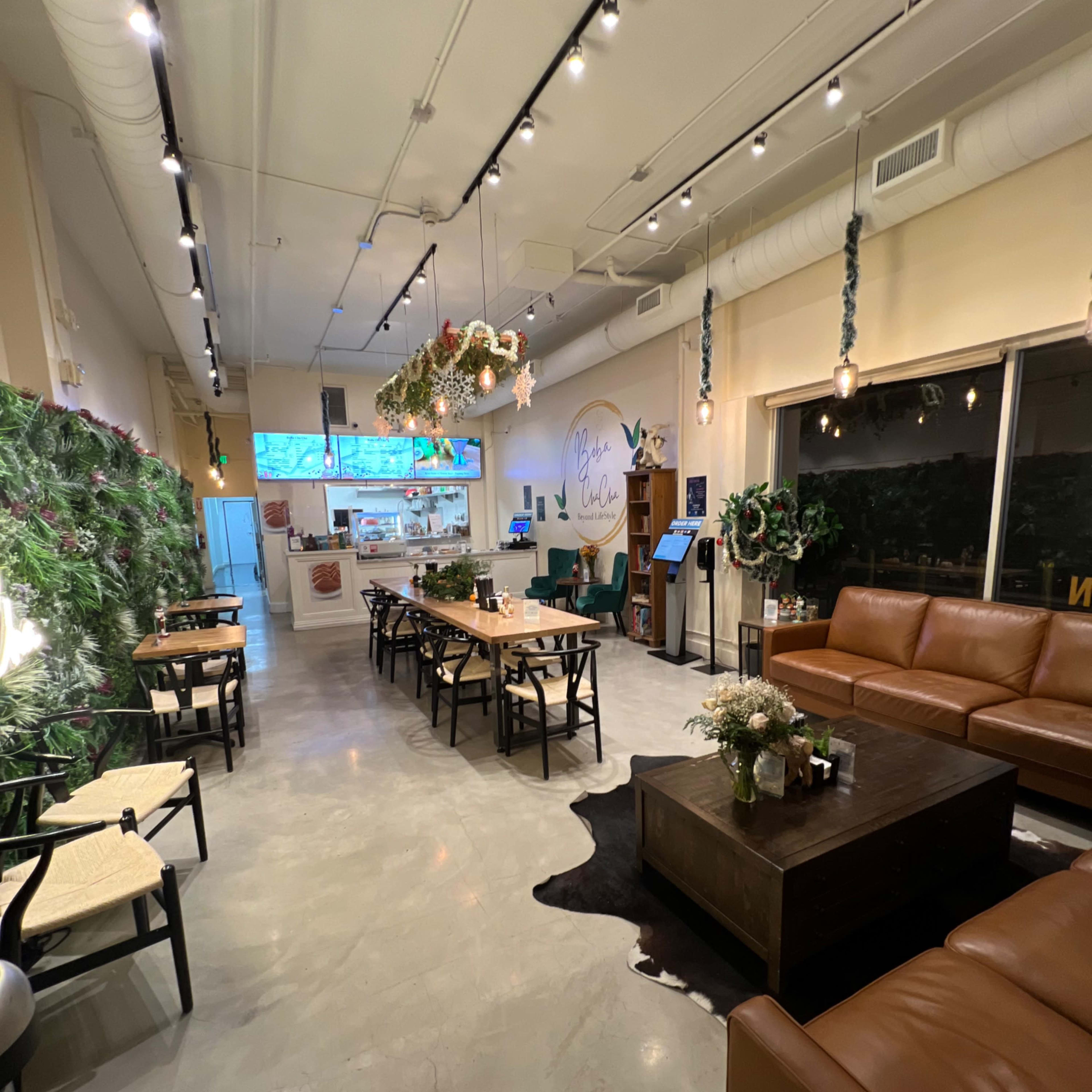 A spacious cafe interior with a mix of seating arrangements, including tables, chairs, and a couch, adorned with plants and decorative elements.