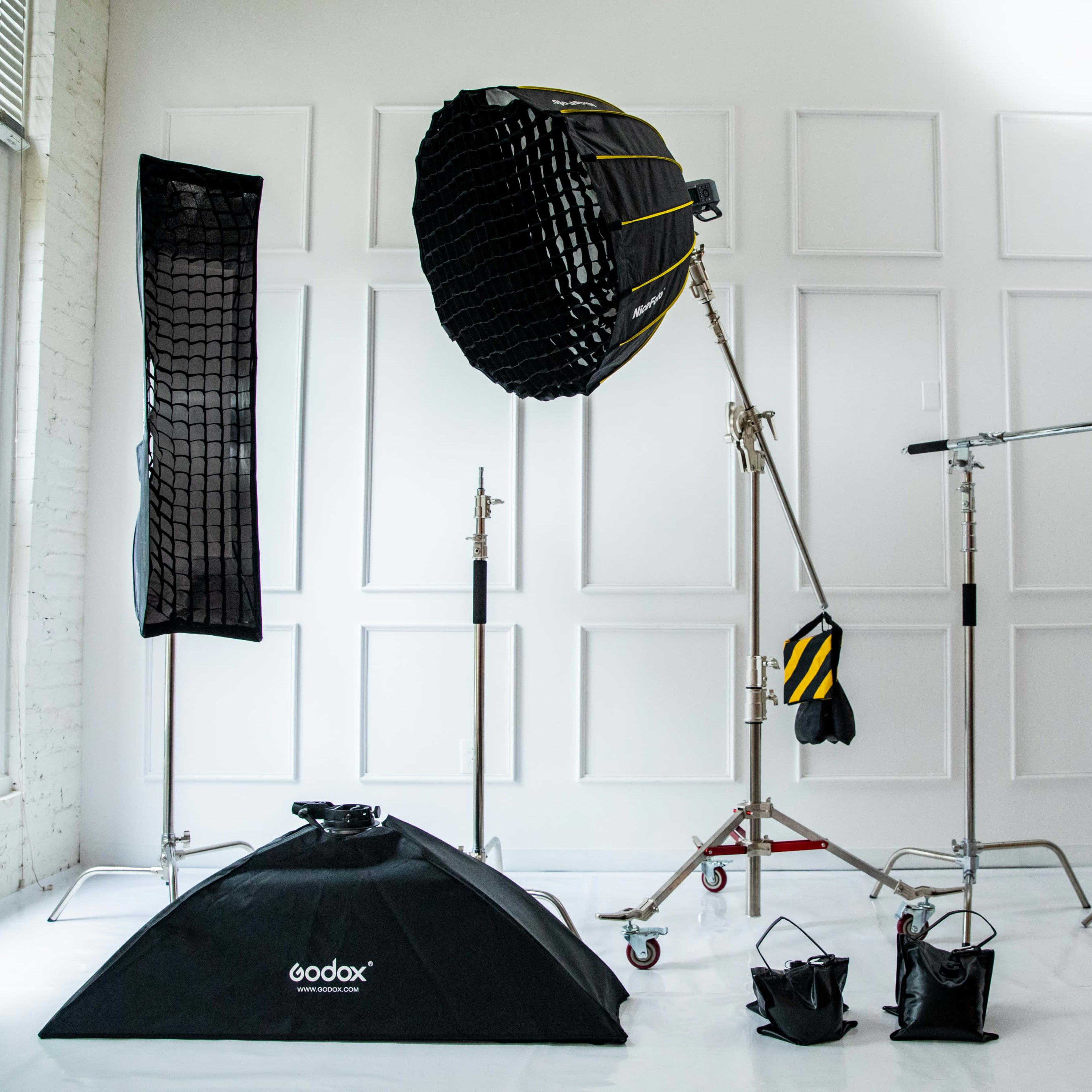 A photography studio setup featuring various lighting equipment, including a large softbox, a light stand, and backdrop supports against a white wall.