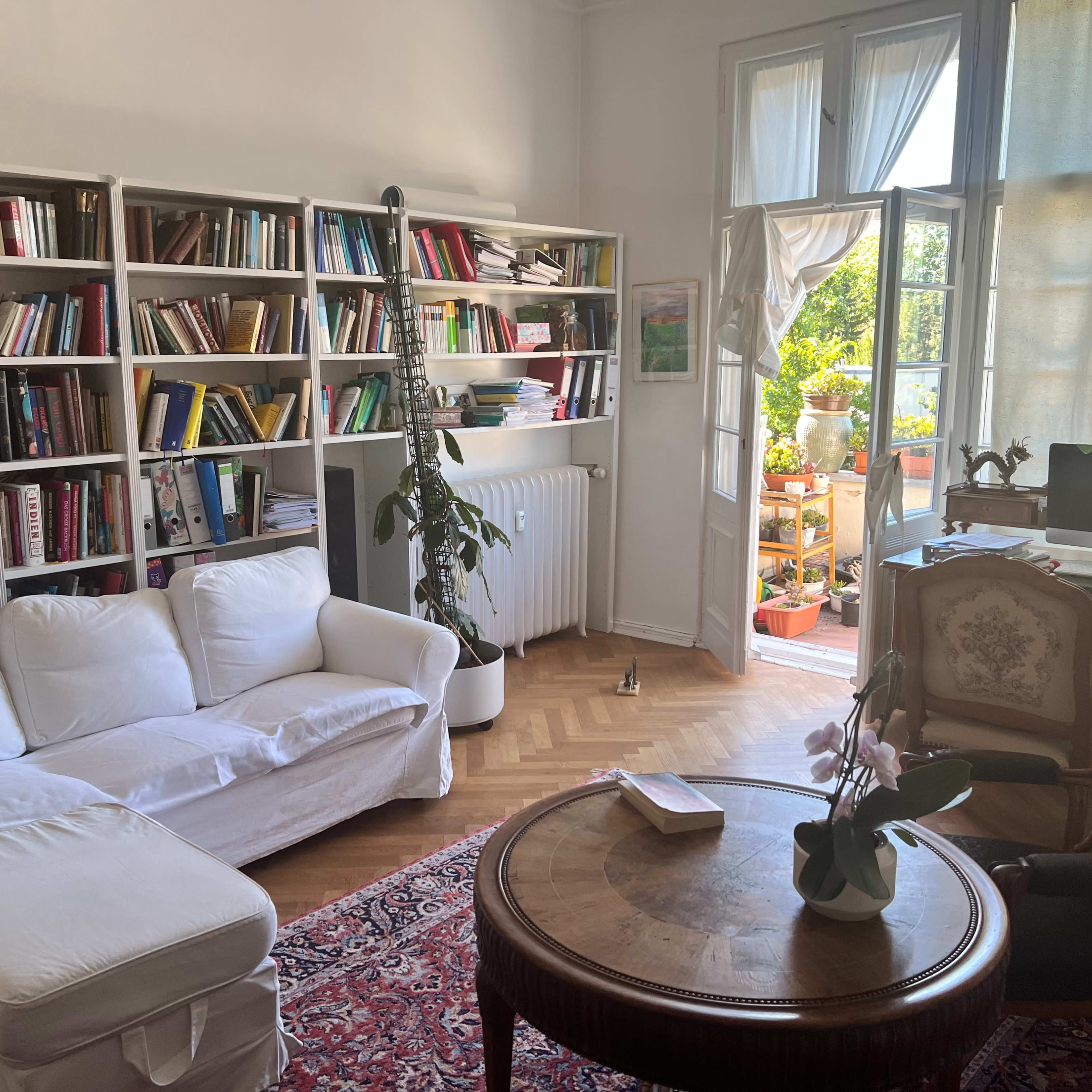A cozy living room features a white sofa, a round wooden table, bookshelves filled with books, and a bright view of a balcony with plants.