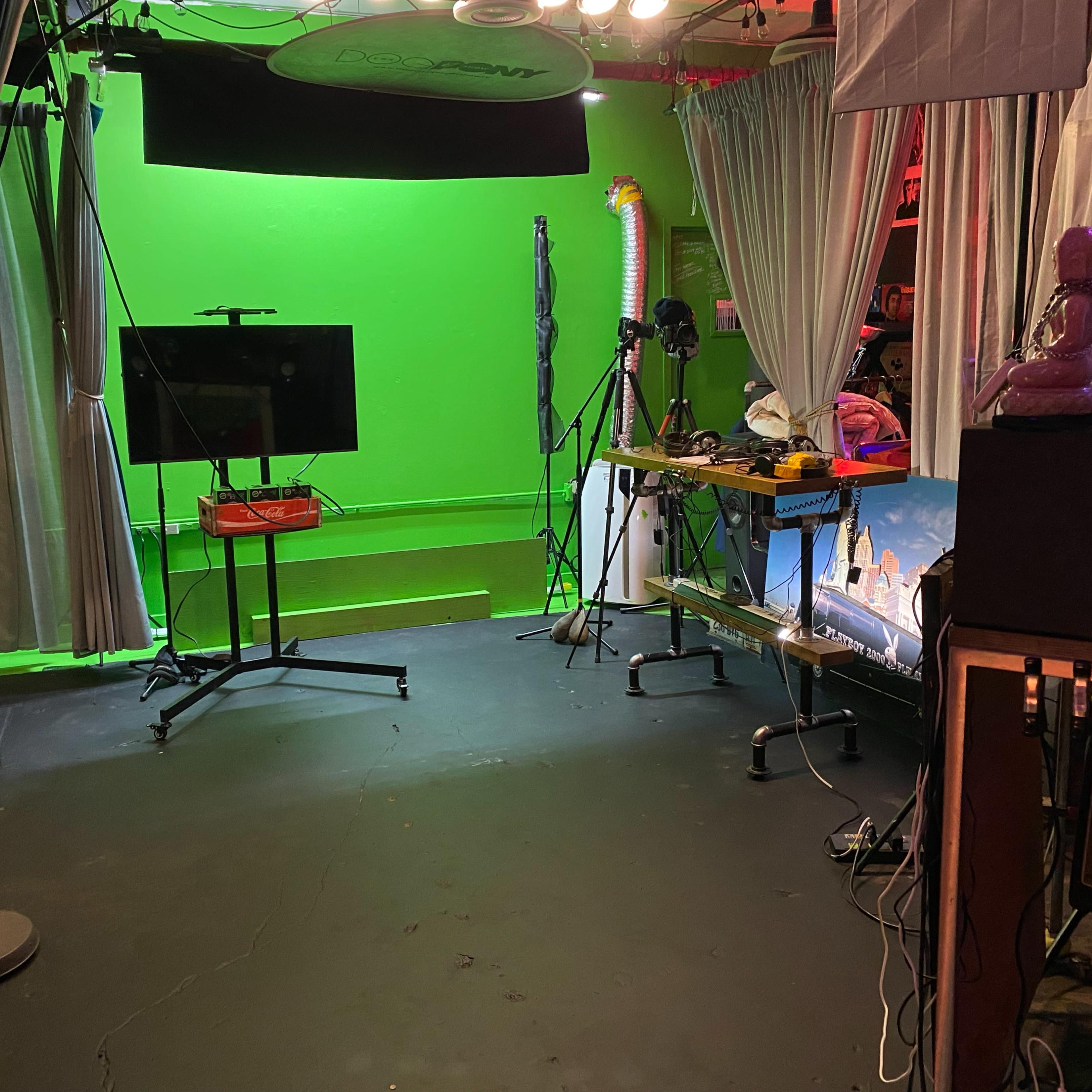 A studio space with a green screen, a television on a stand, various equipment, and draped curtains.
