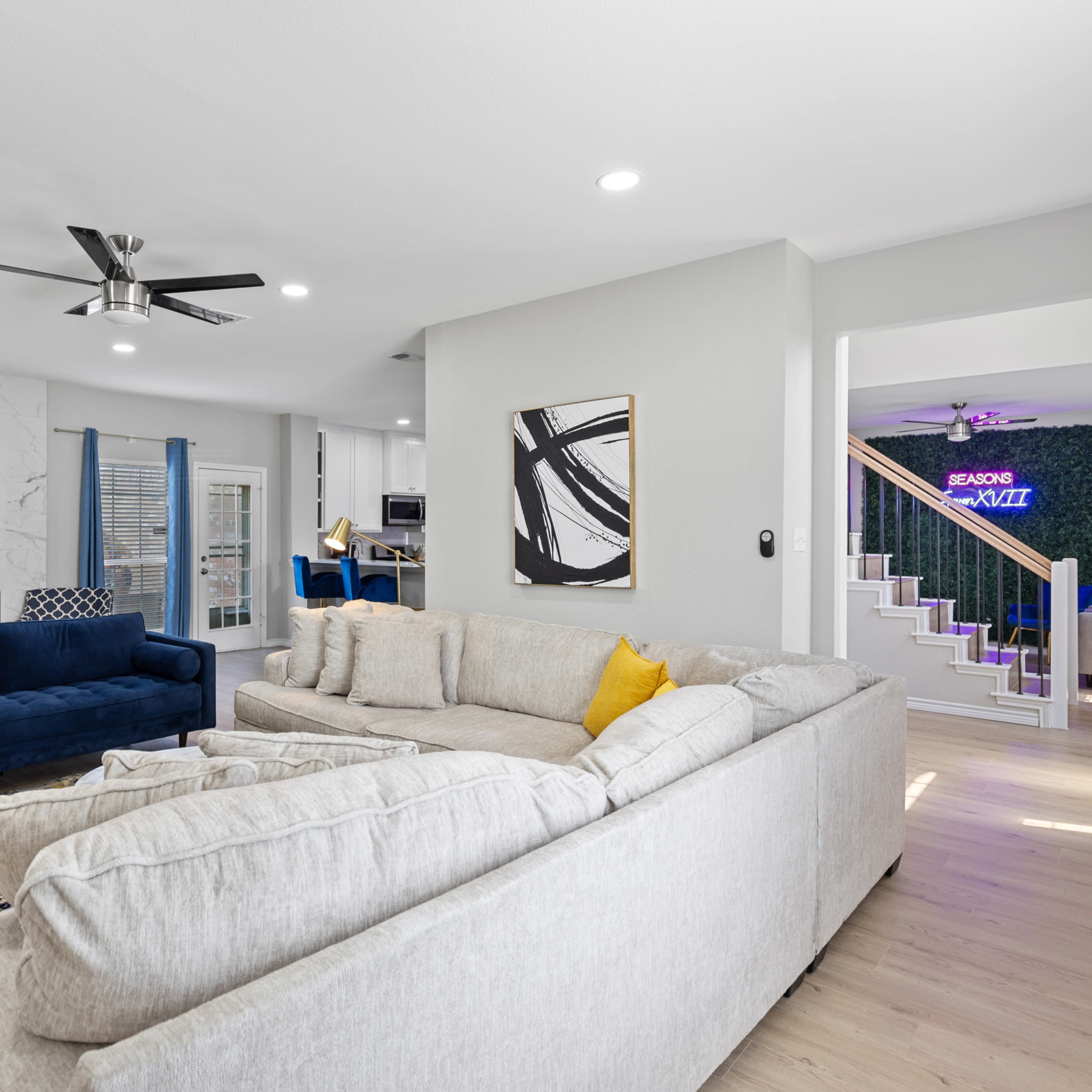 A modern living room with a blue sofa and a light-colored sectional, set against a backdrop of a marble accent wall and a staircase, with a planted space visible in the background.
