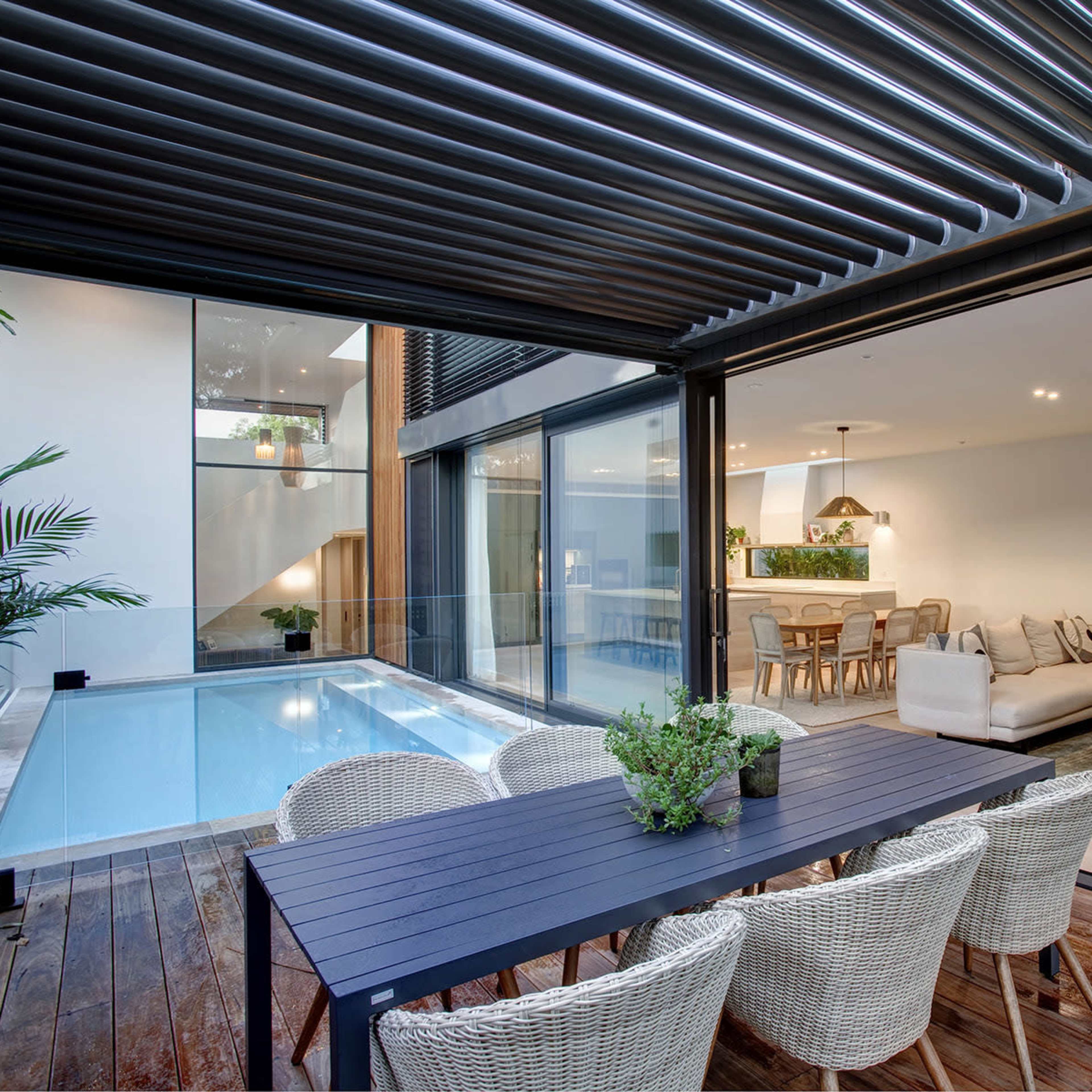 A modern indoor-outdoor living space featuring a dining area with a table and chairs next to a swimming pool, with large glass walls that open into a stylish living room.