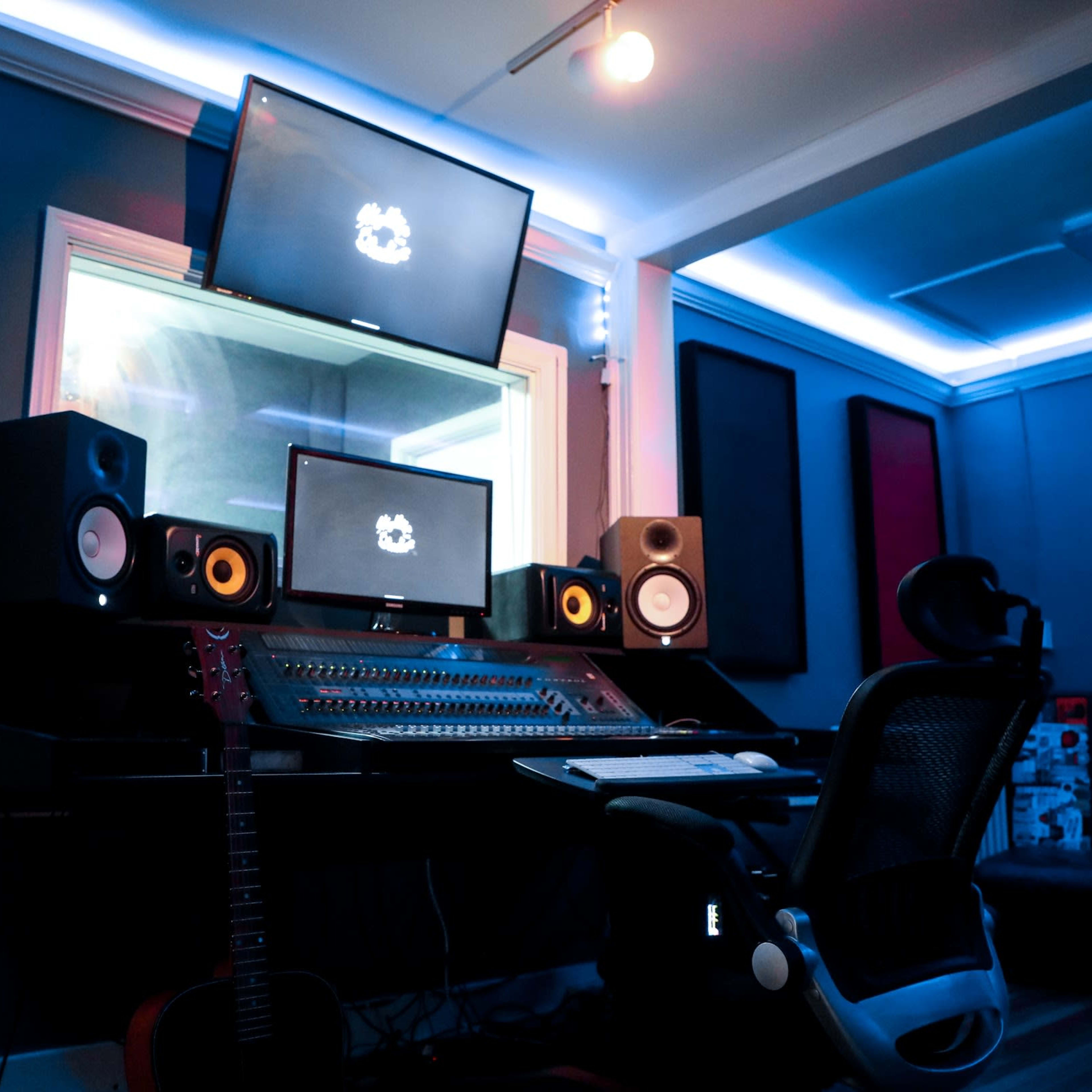 A music studio is equipped with multiple monitors, speakers, a mixing console, and a chair, all illuminated by blue LED lighting.