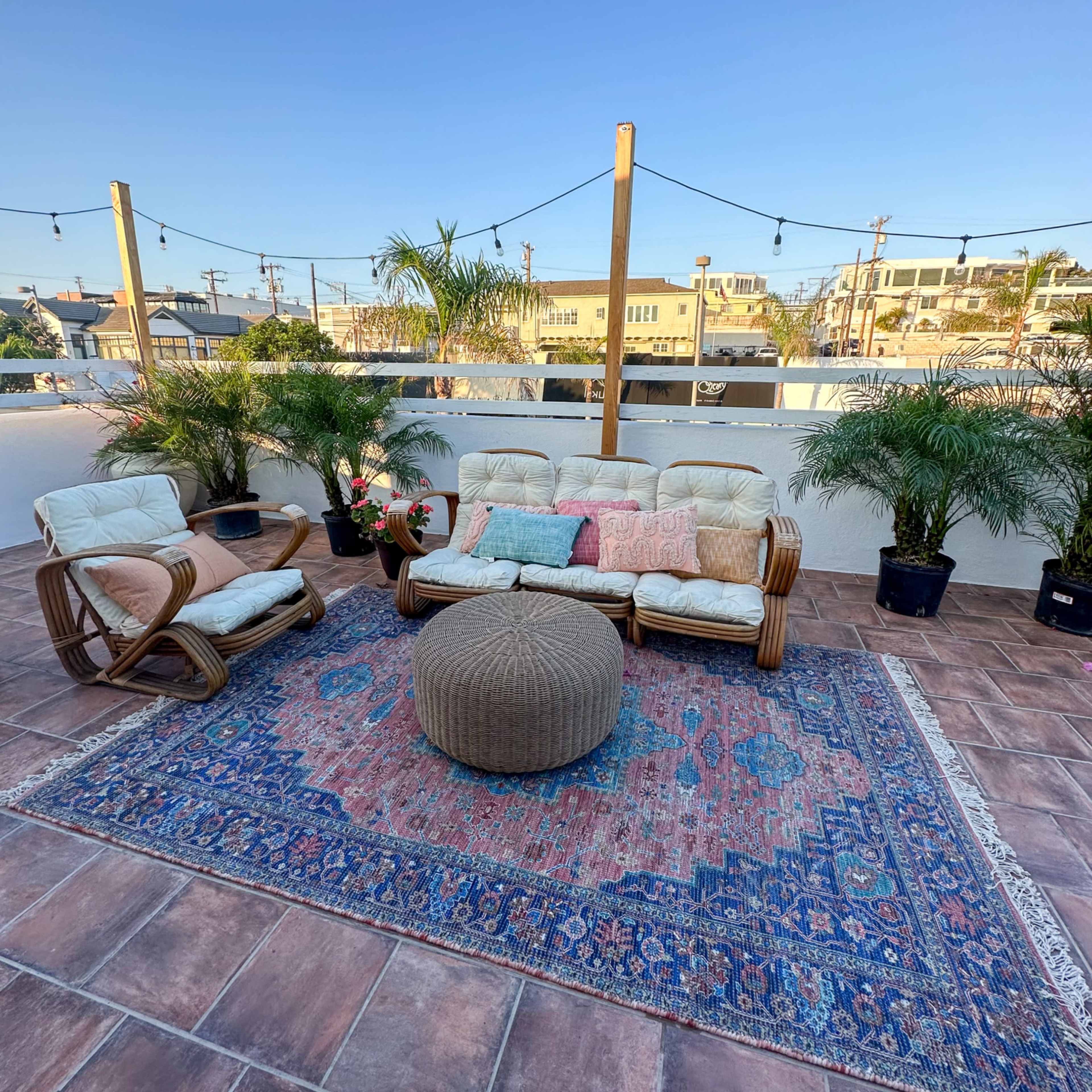 A rooftop seating area features two chairs and a couch on a patterned rug, surrounded by potted plants and with a clear blue sky above.