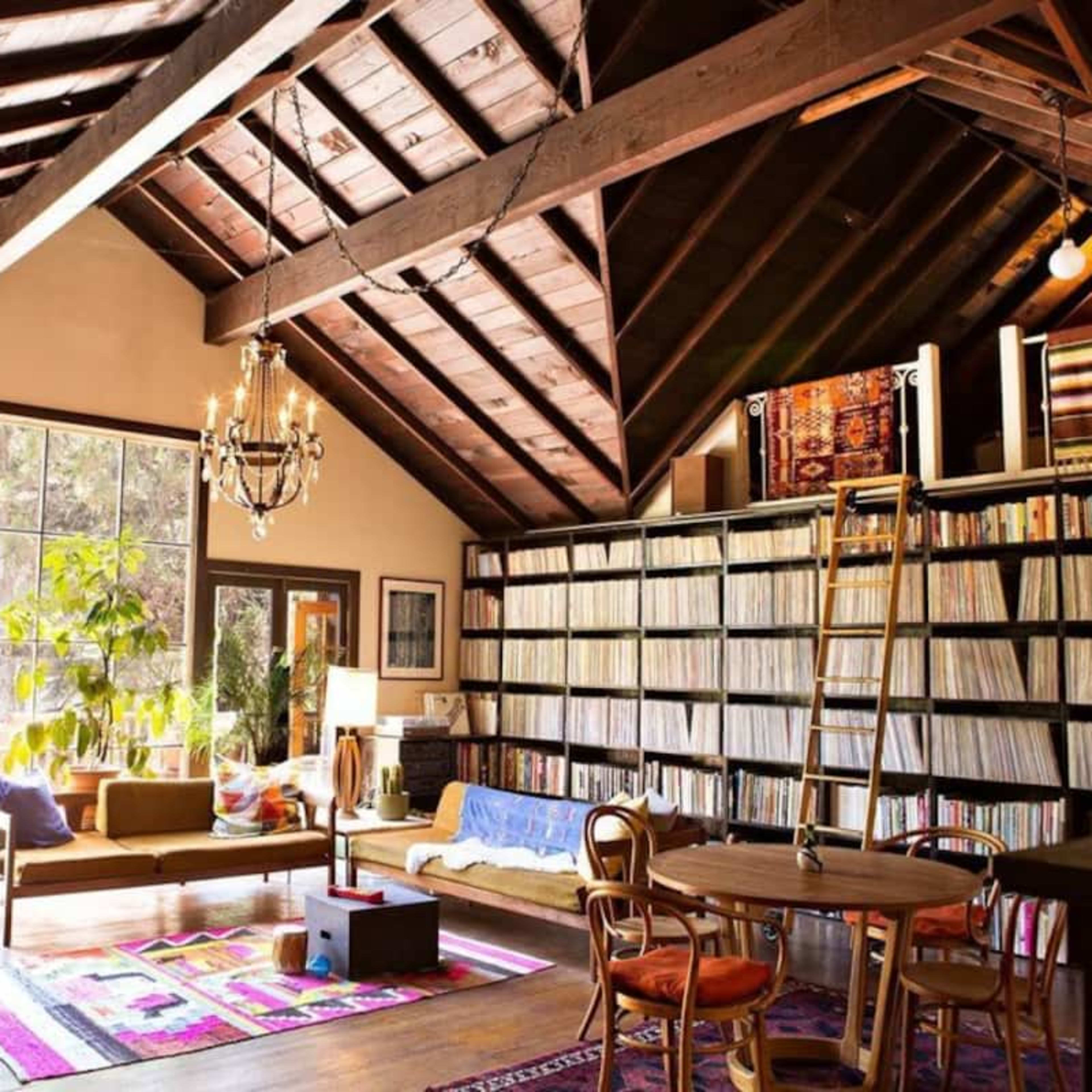 A spacious living room with high wooden ceilings, a large bookshelf filled with records, and comfortable seating area beside large windows.