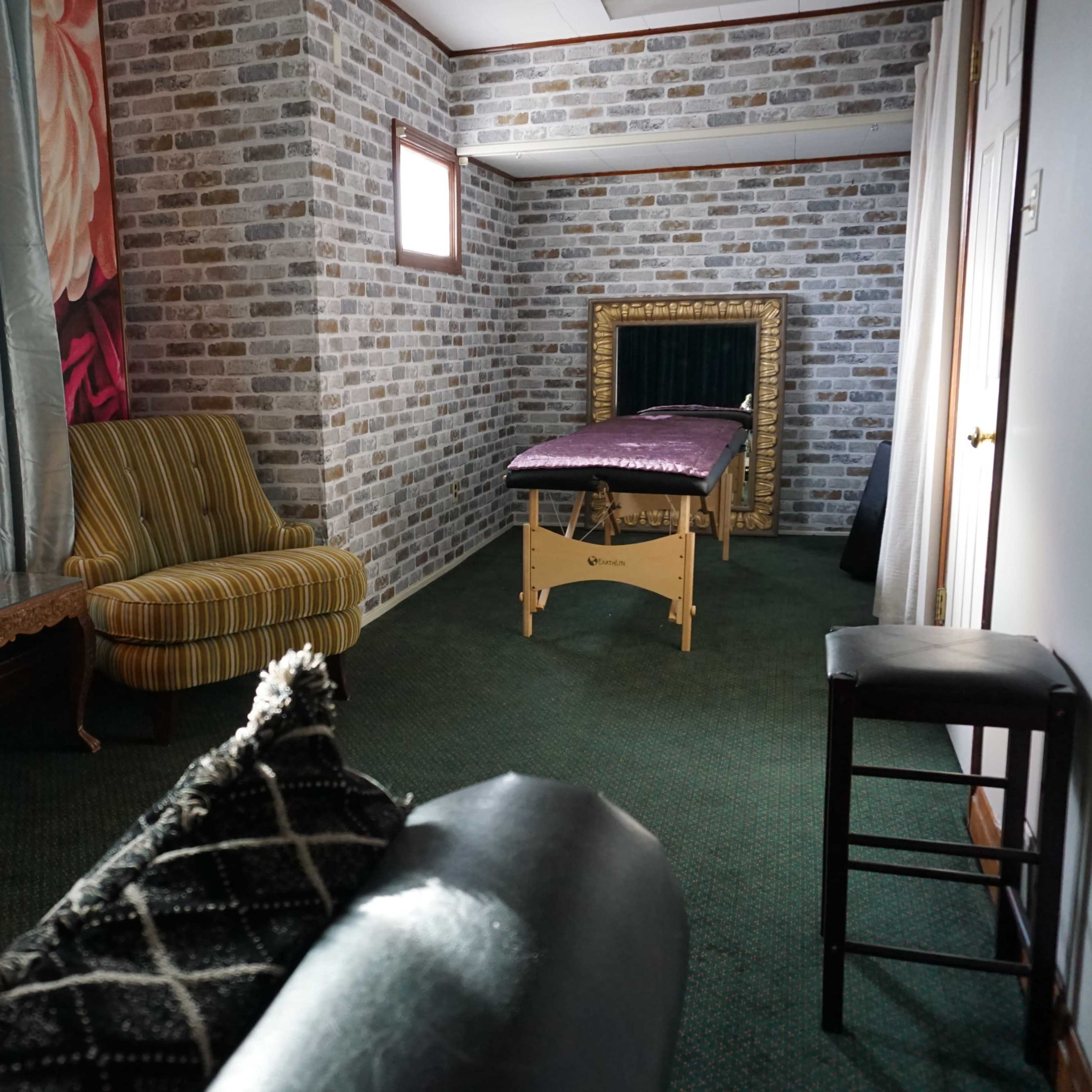 A room with a massage table against a brick-patterned wall, a striped armchair, a wooden table, and a stool, all illuminated by a standing lamp.