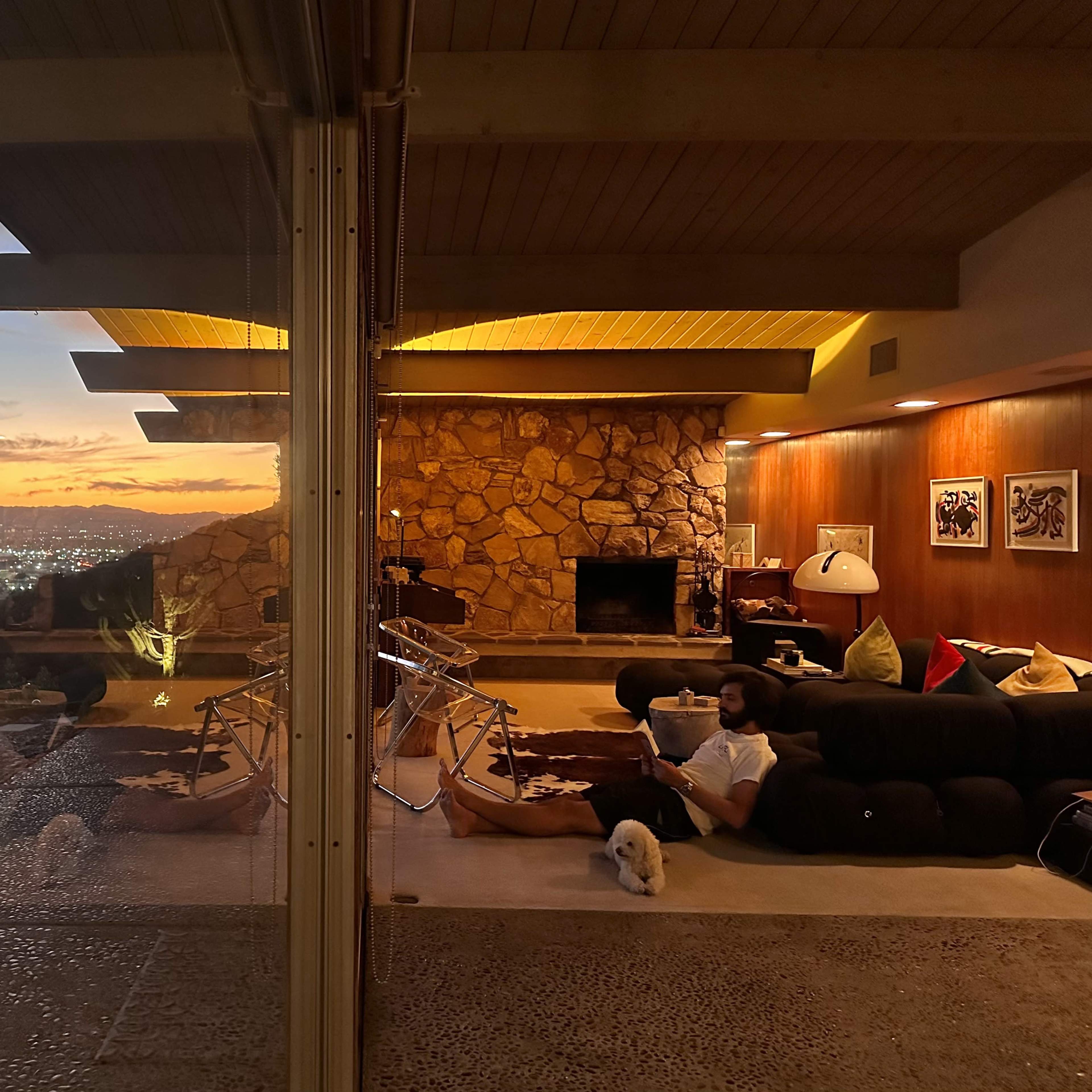A person sits on a couch with a small dog in a modern living room that opens to a view of a sunset over a city landscape.