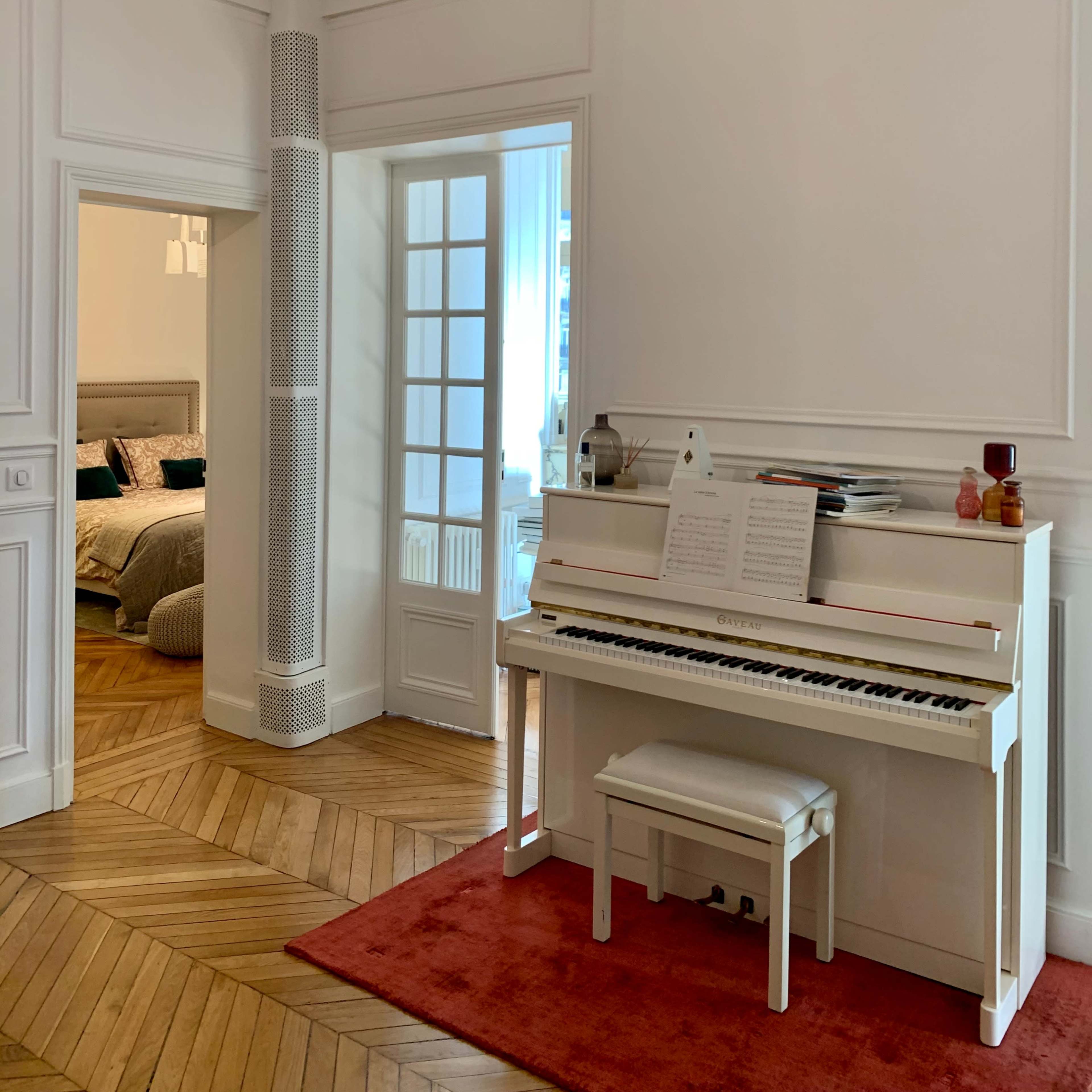 A white piano is positioned against a wall in a room with wooden flooring, leading into a bedroom through a glass-paned door.