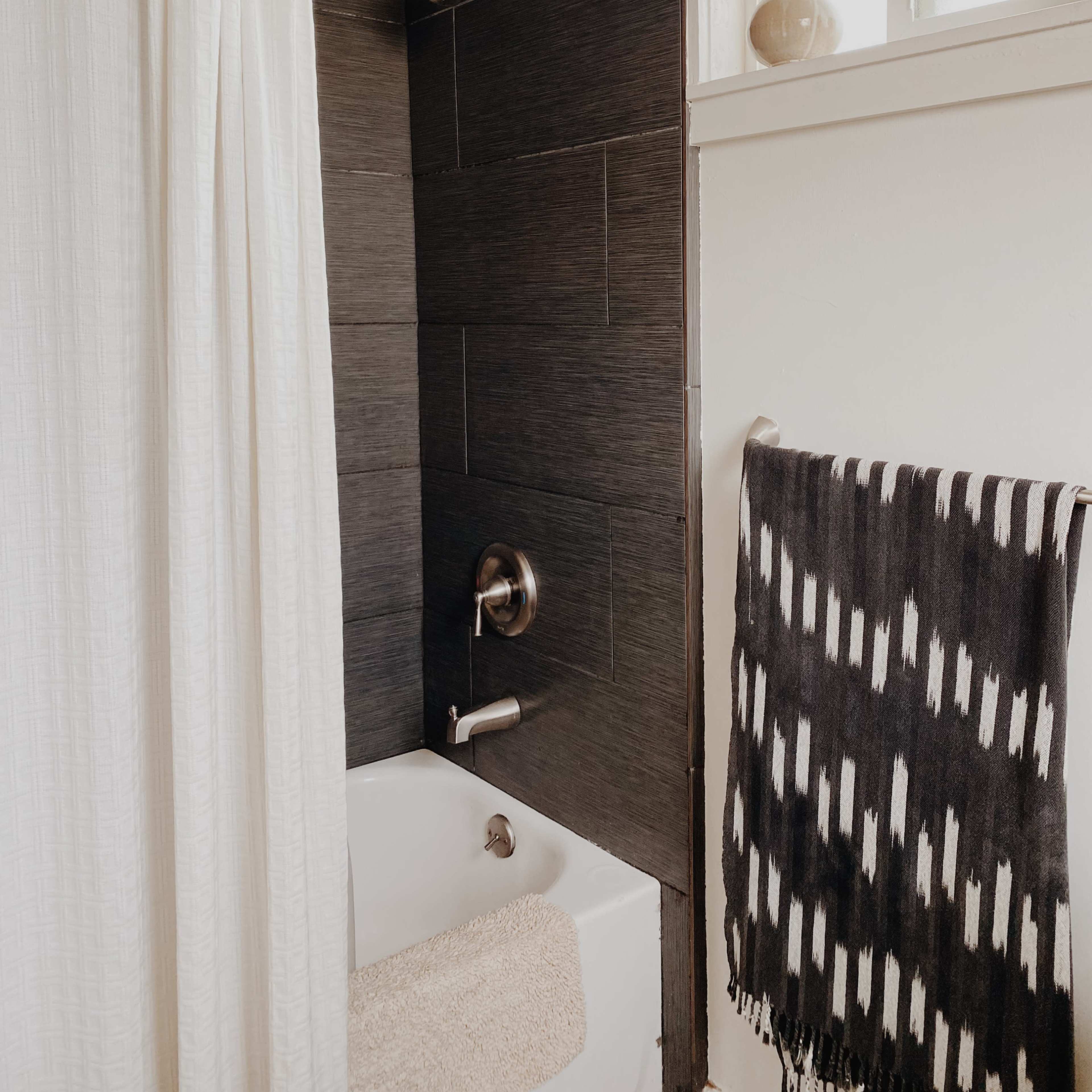 A corner of a bathroom featuring a white shower curtain, a black-tiled wall, a bathtub with a faucet, and a towel hanging on a rack.