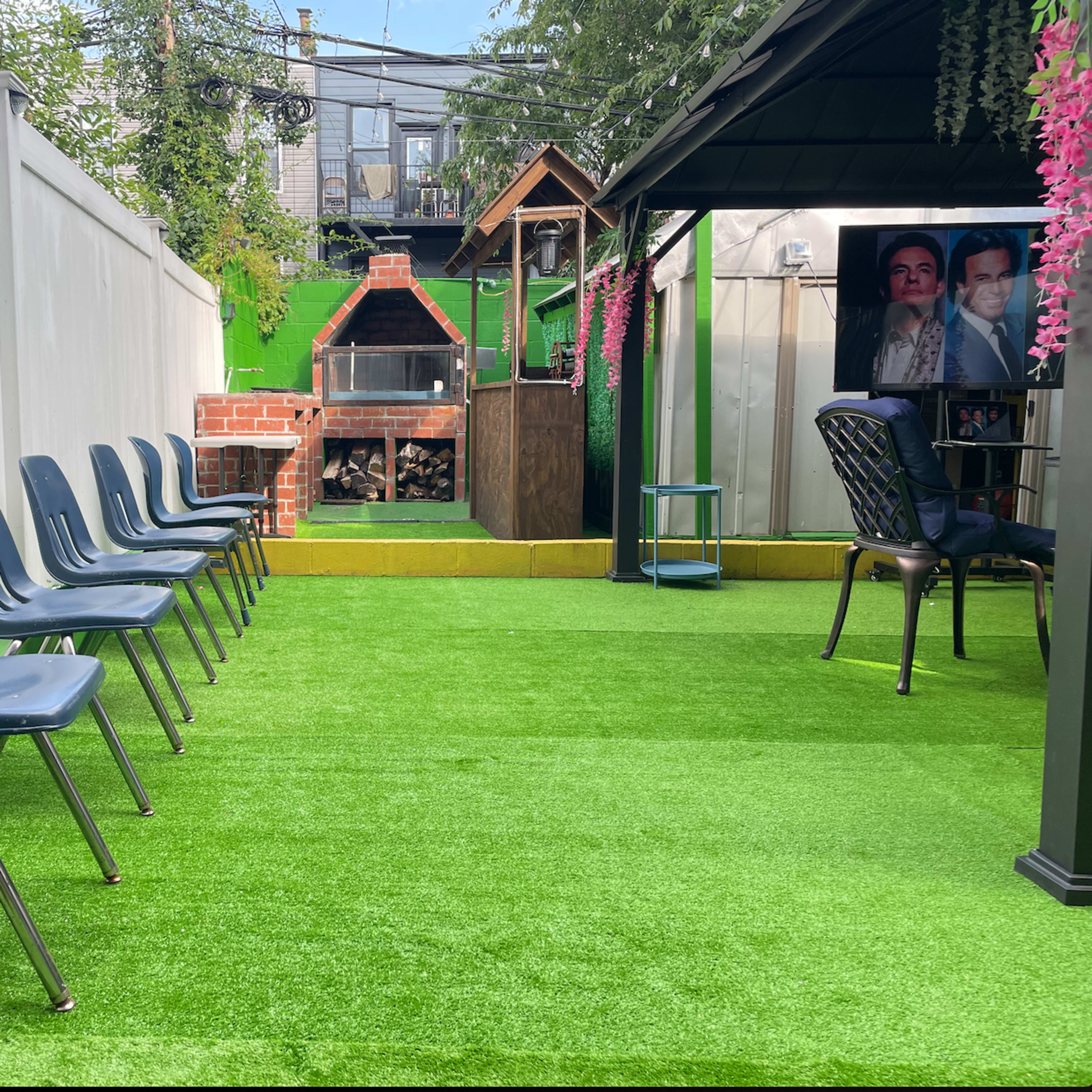 An outdoor space with a row of blue chairs on artificial grass, a barbecue area in the background, and a small gazebo with portraits visible.