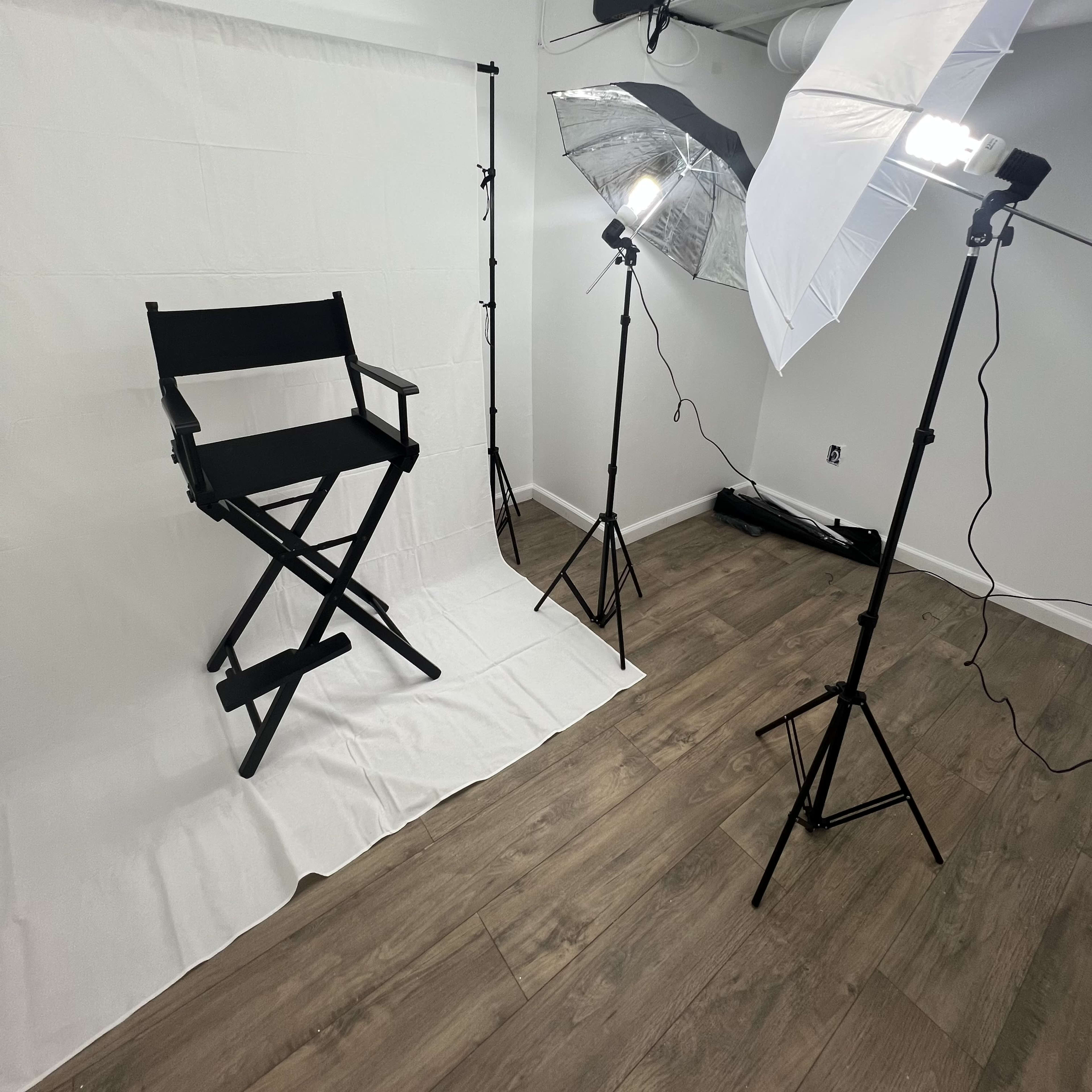 A photography setup with a black director's chair on a white backdrop, flanked by two light stands with softboxes, positioned on a wooden floor.