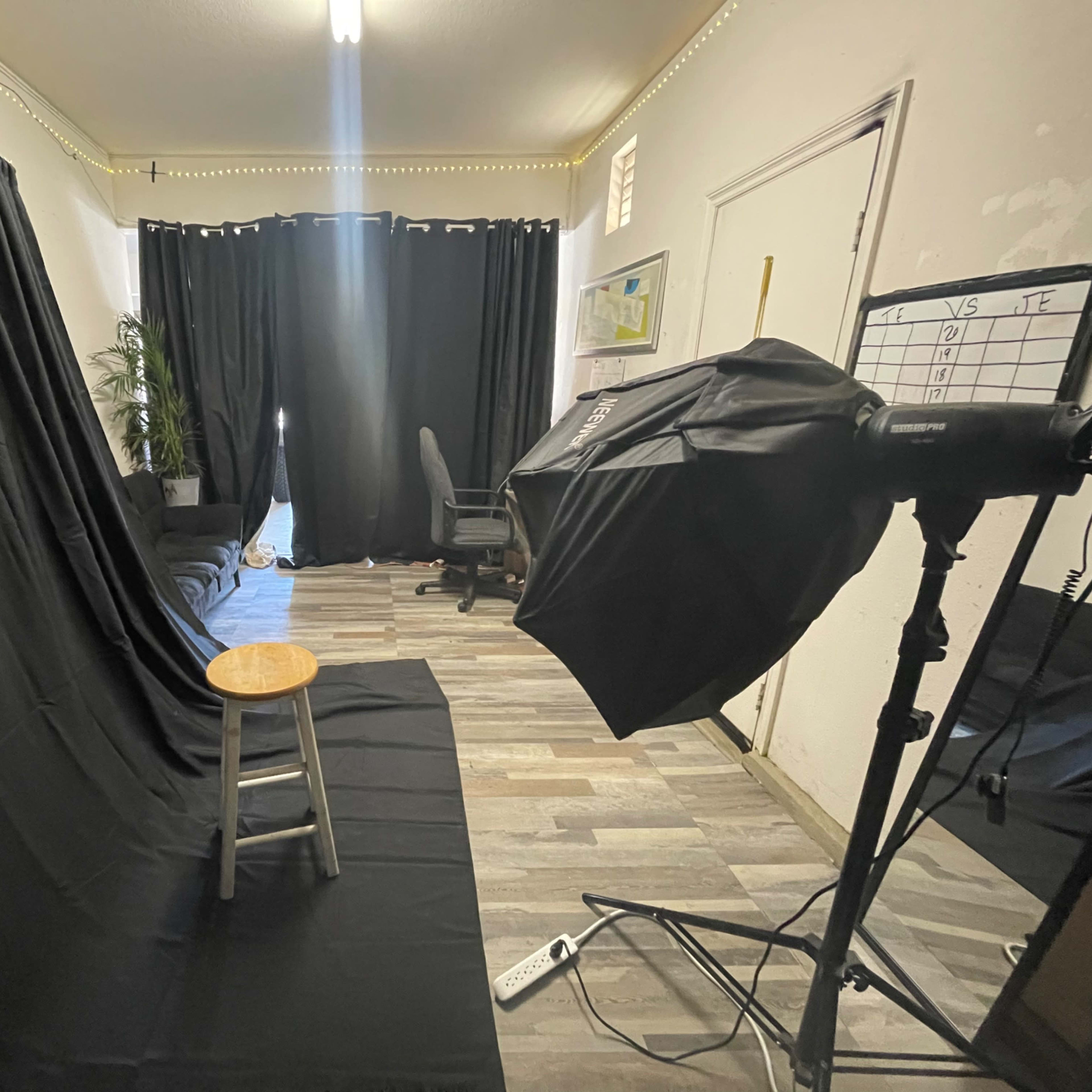 A photography setup with a black backdrop, a wooden stool in the center, and a lighting equipment on the side, all located in a room with dark curtains and minimal furniture.