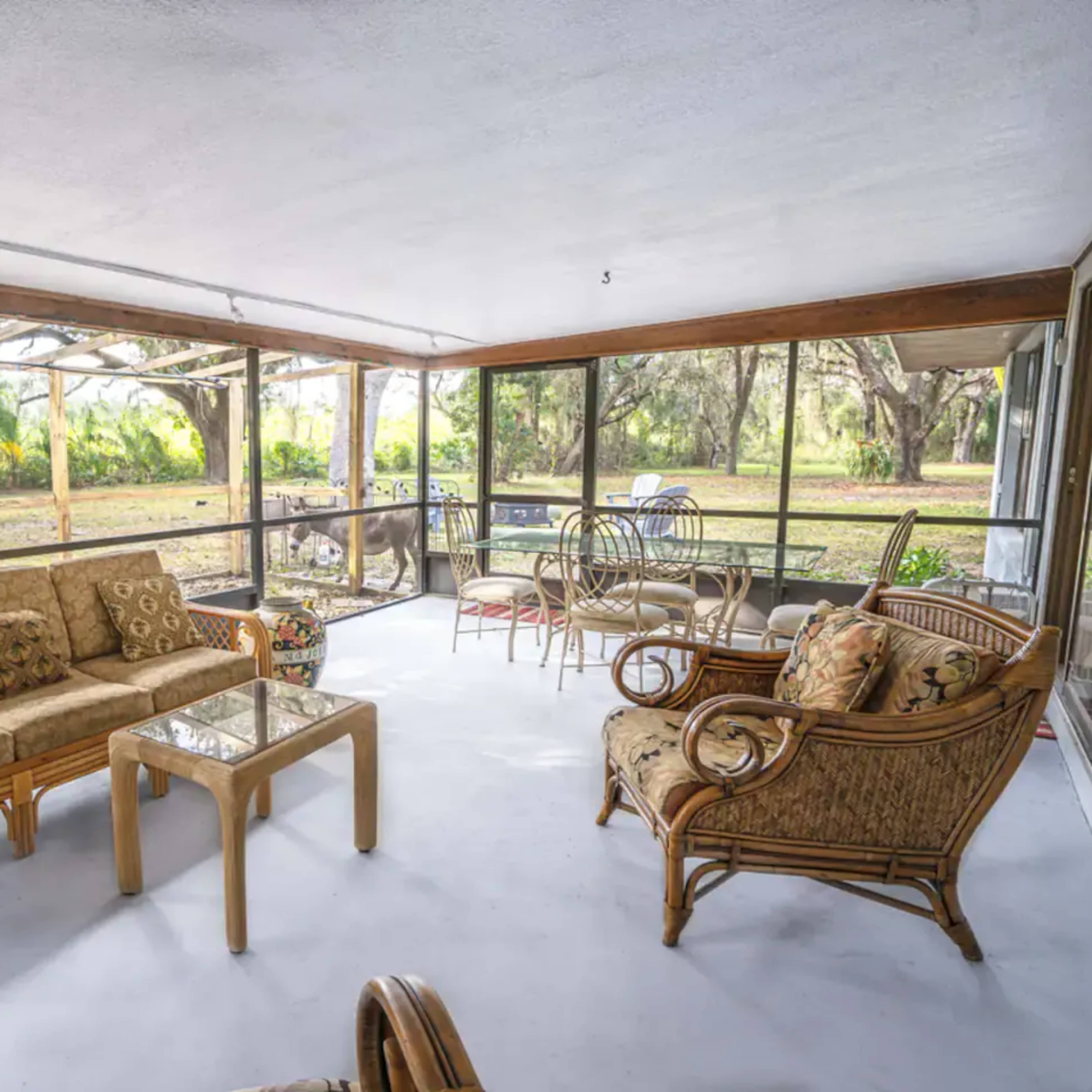A sunroom with wicker furniture, including a sofa, armchairs, and a glass coffee table, overlooking a green outdoor area through large screened windows.