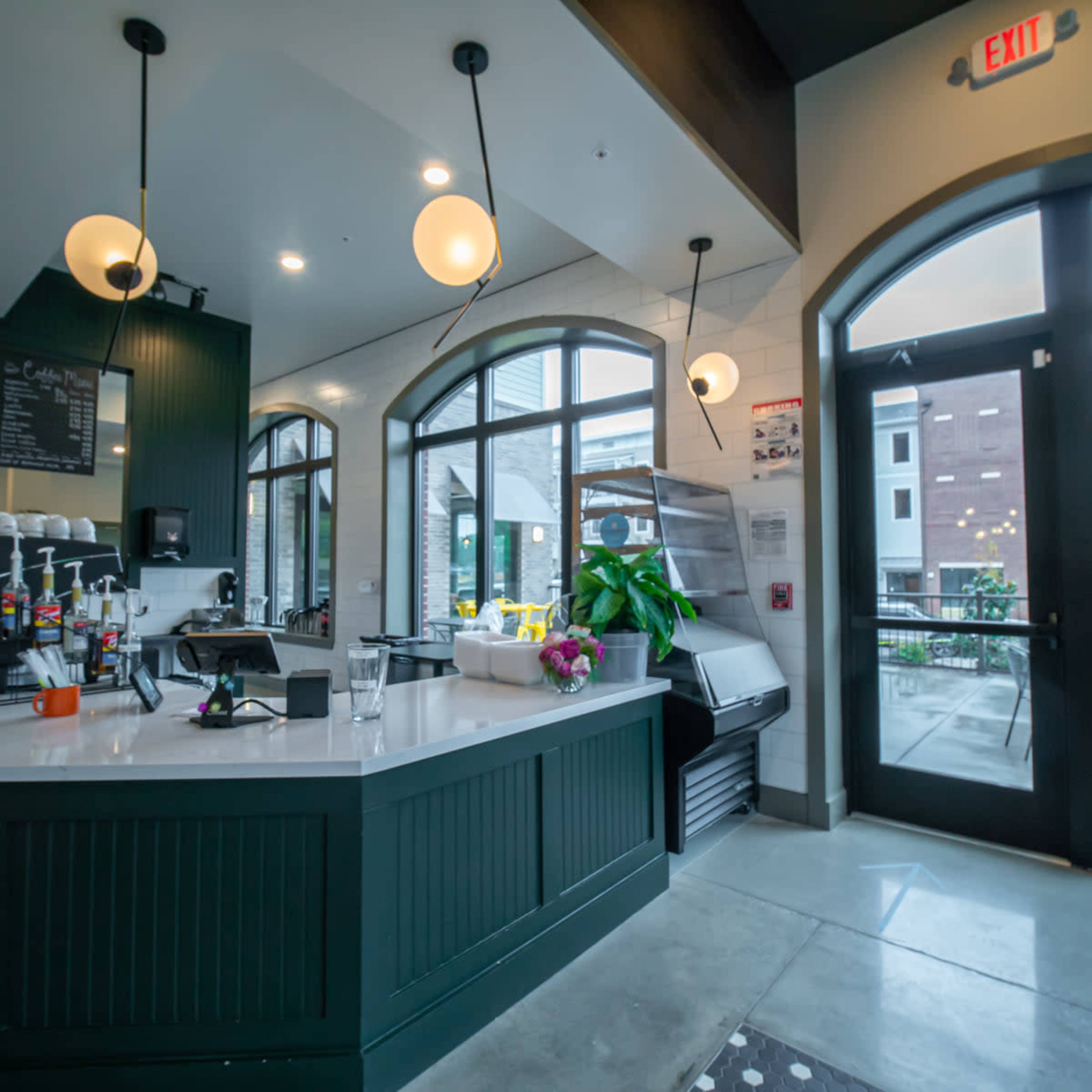 A modern coffee shop interior with a white countertop, green cabinetry, and large arched windows that open to an outdoor seating area.