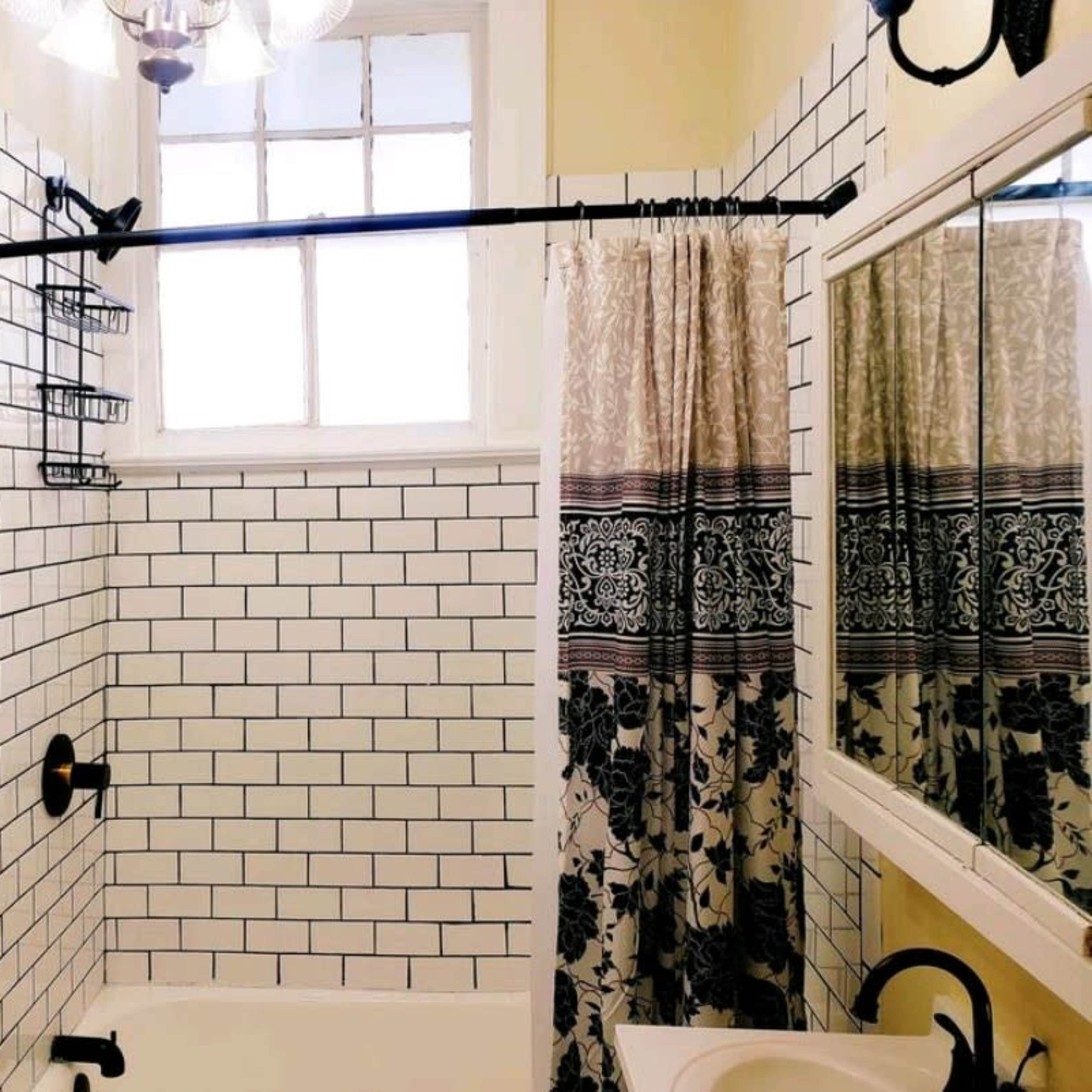 A bright bathroom features white tiled walls, a tub with a black faucet, and a window with a decorative curtain.