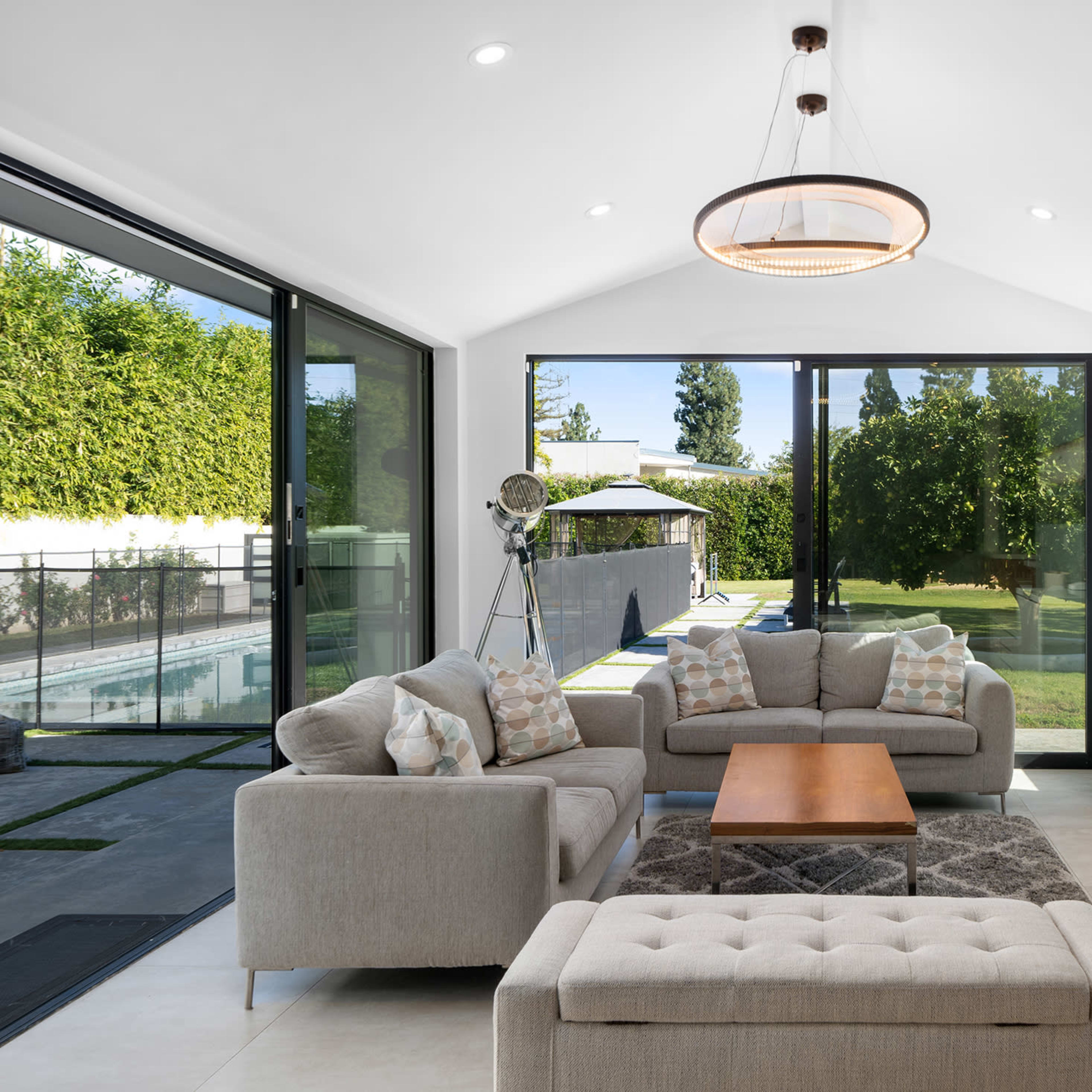 The living room features a light-colored sofa set, a wooden coffee table, and large windows offering a view of the backyard and pool area.