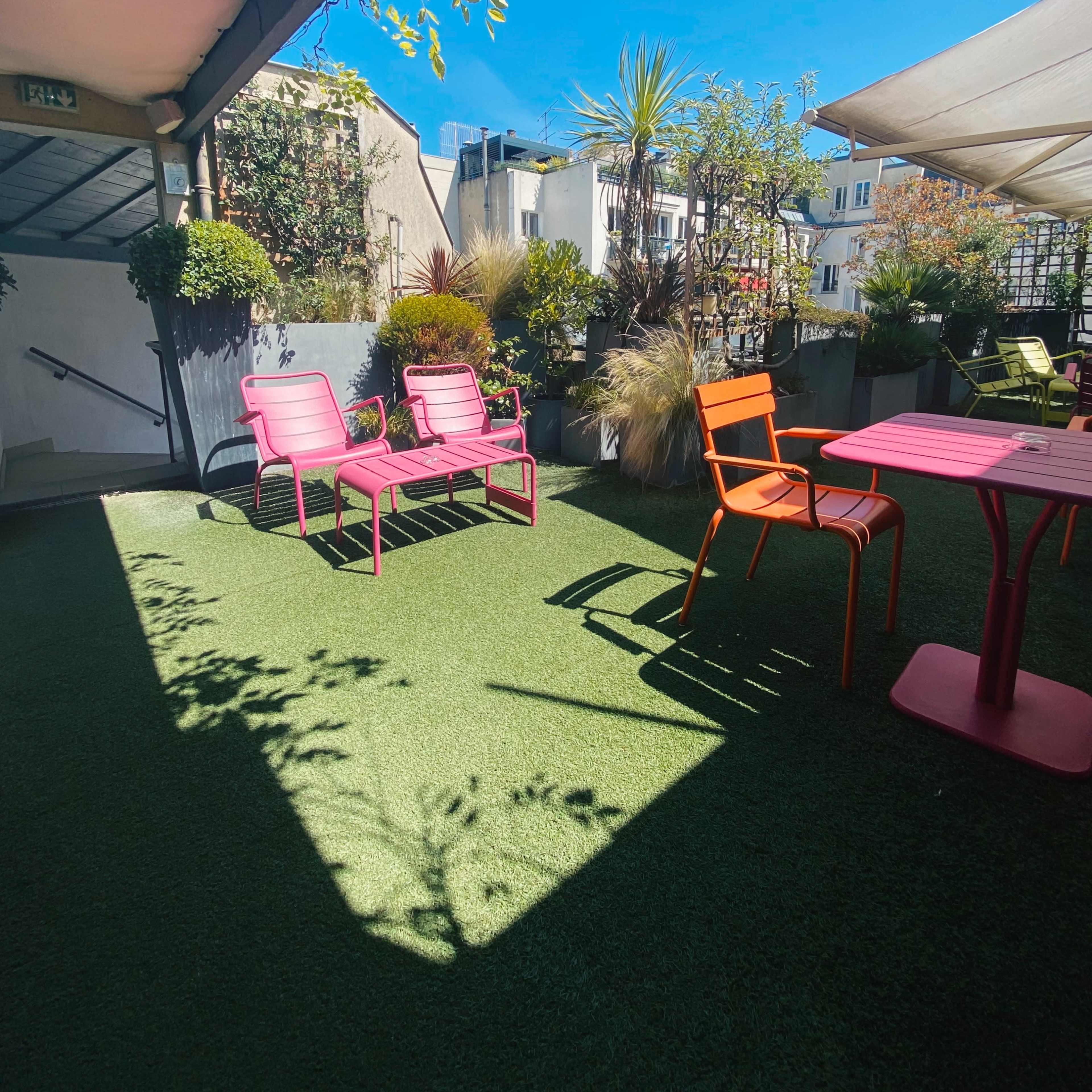 A rooftop terrace with pink chairs and a table surrounded by greenery under a clear blue sky.
