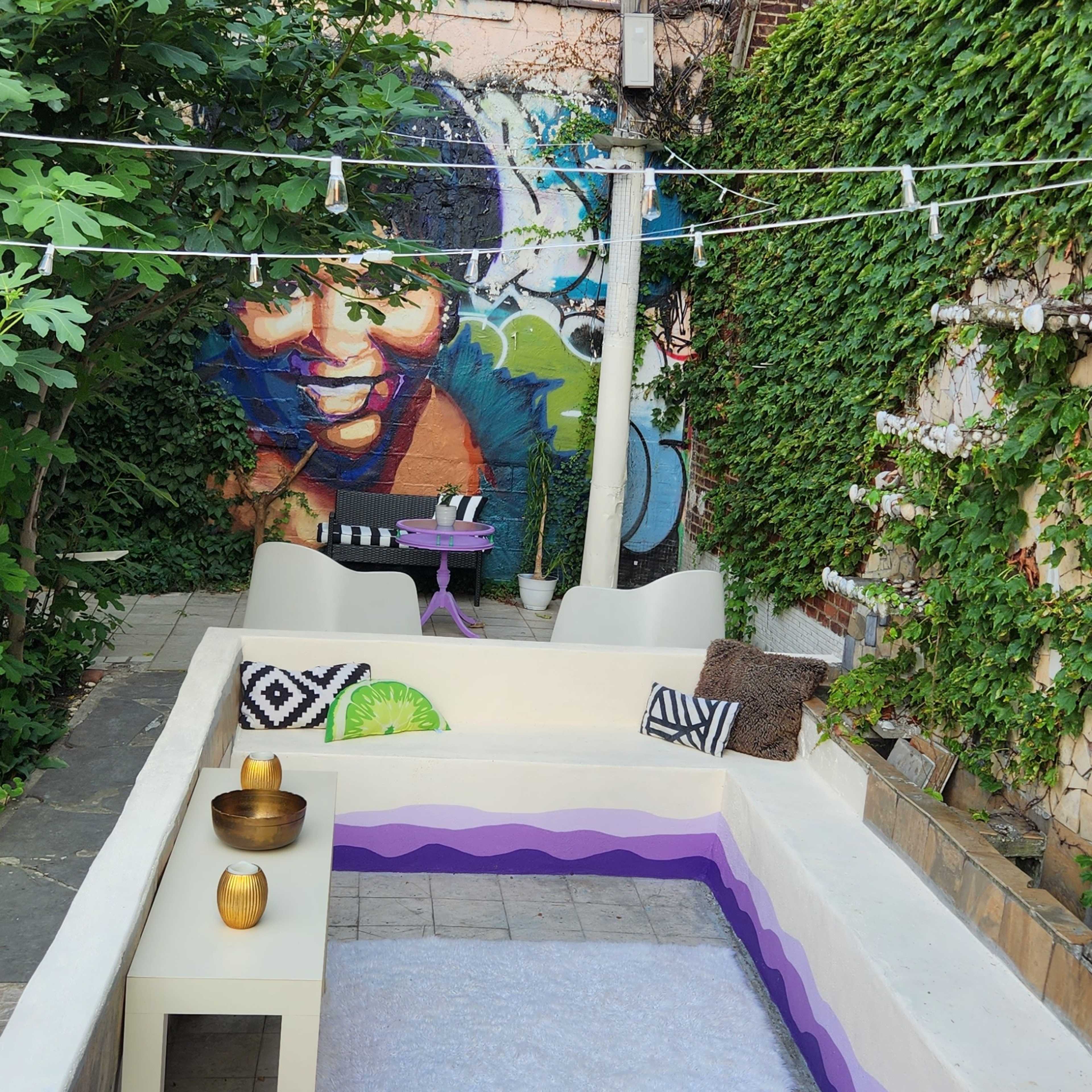 A small outdoor courtyard features a white lounging area, colorful cushions, and a vibrant mural on the brick wall.