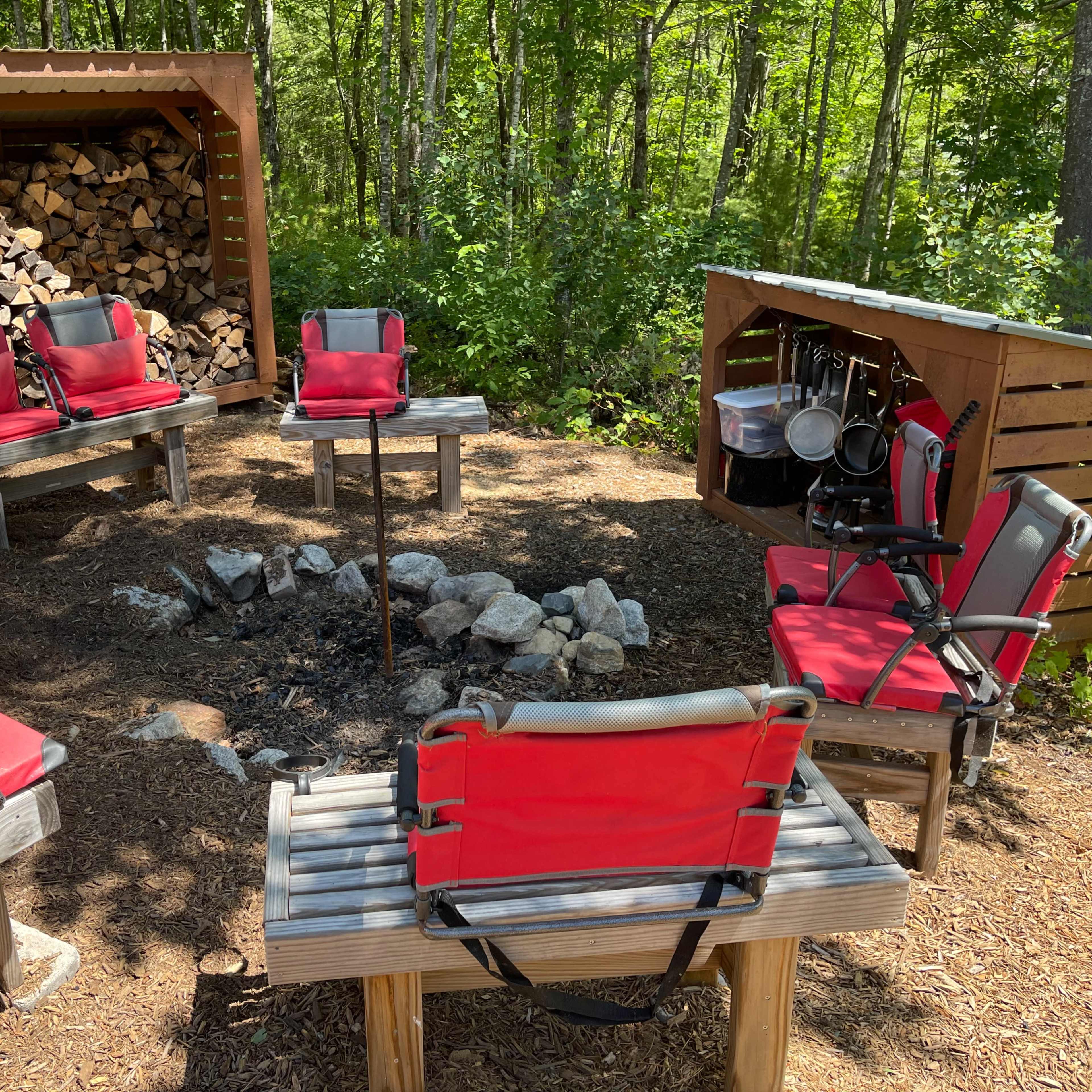 A clearing in the woods features a circular arrangement of red camping chairs around a fire pit, alongside a storage shed for firewood and cooking equipment.