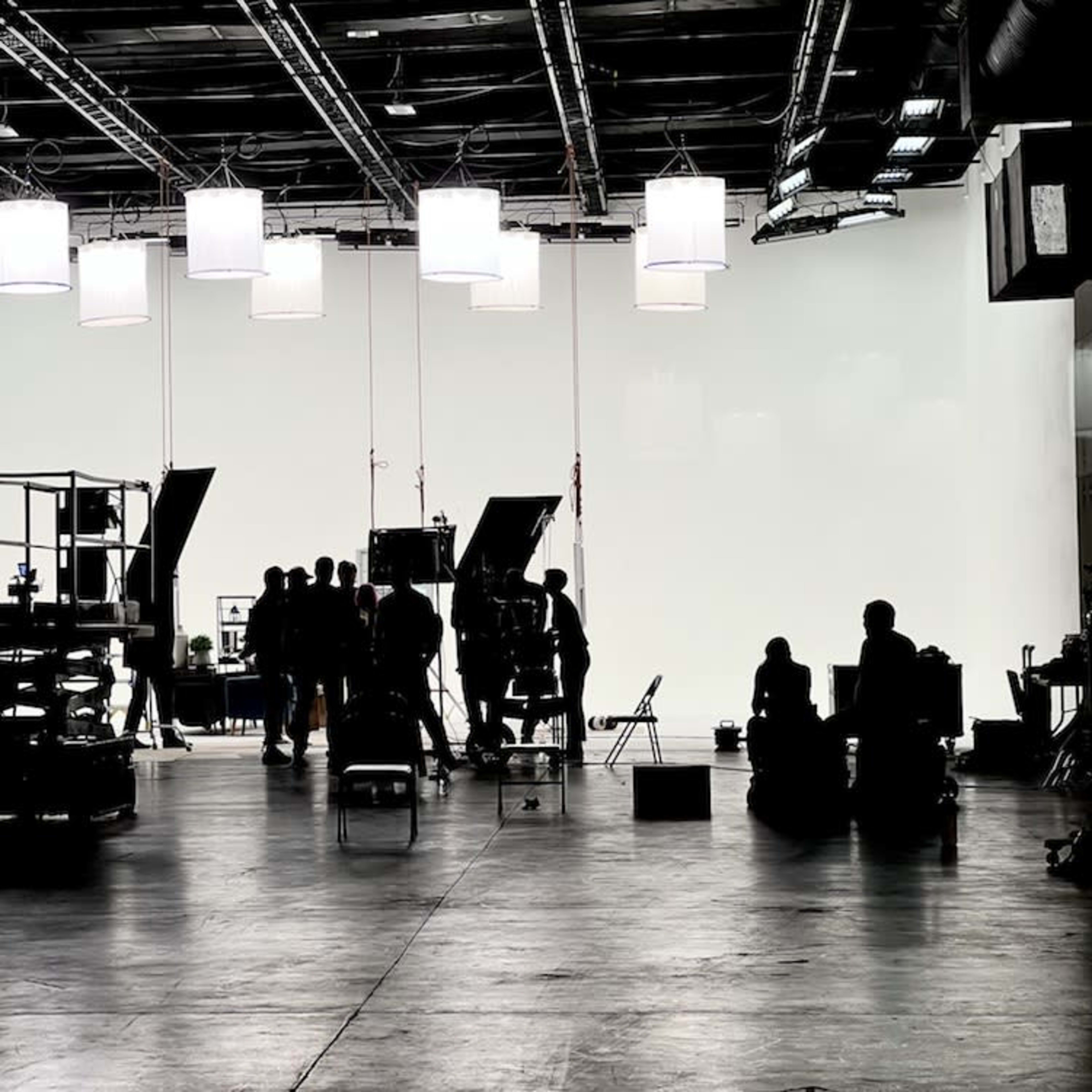 A film set is illuminated by overhead lights, featuring various equipment and crew members working in the foreground.